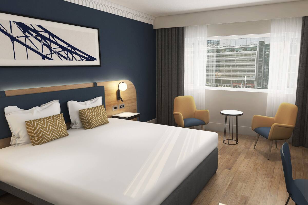 A hotel room with a large bed, two armchairs, a small table, a nightstand with a lamp, a window with curtains, and a cityscape view.