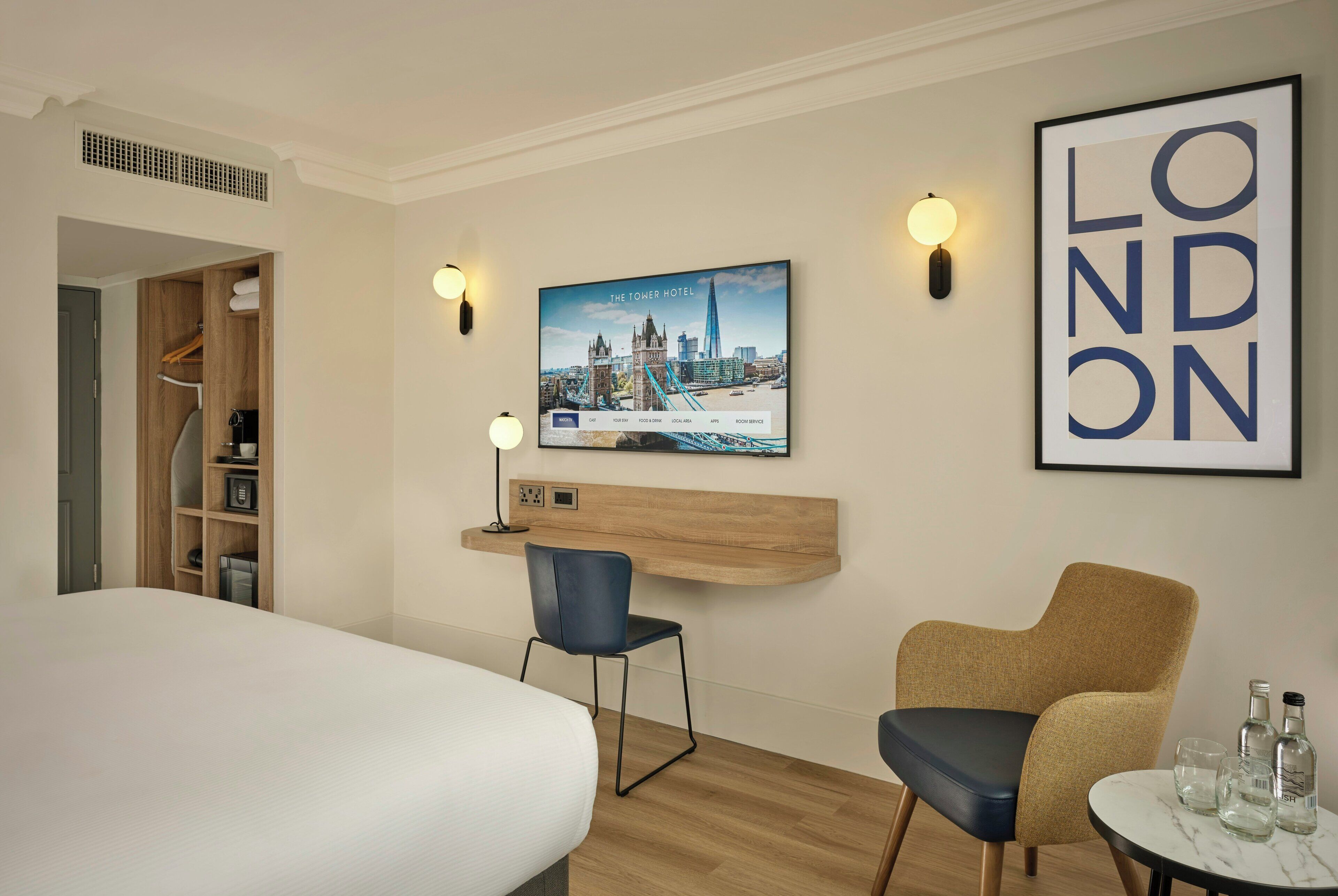 Habitación de hotel con cama, escritorio, silla, televisor montado en la pared y un cuadro con una imagen de Londres.
