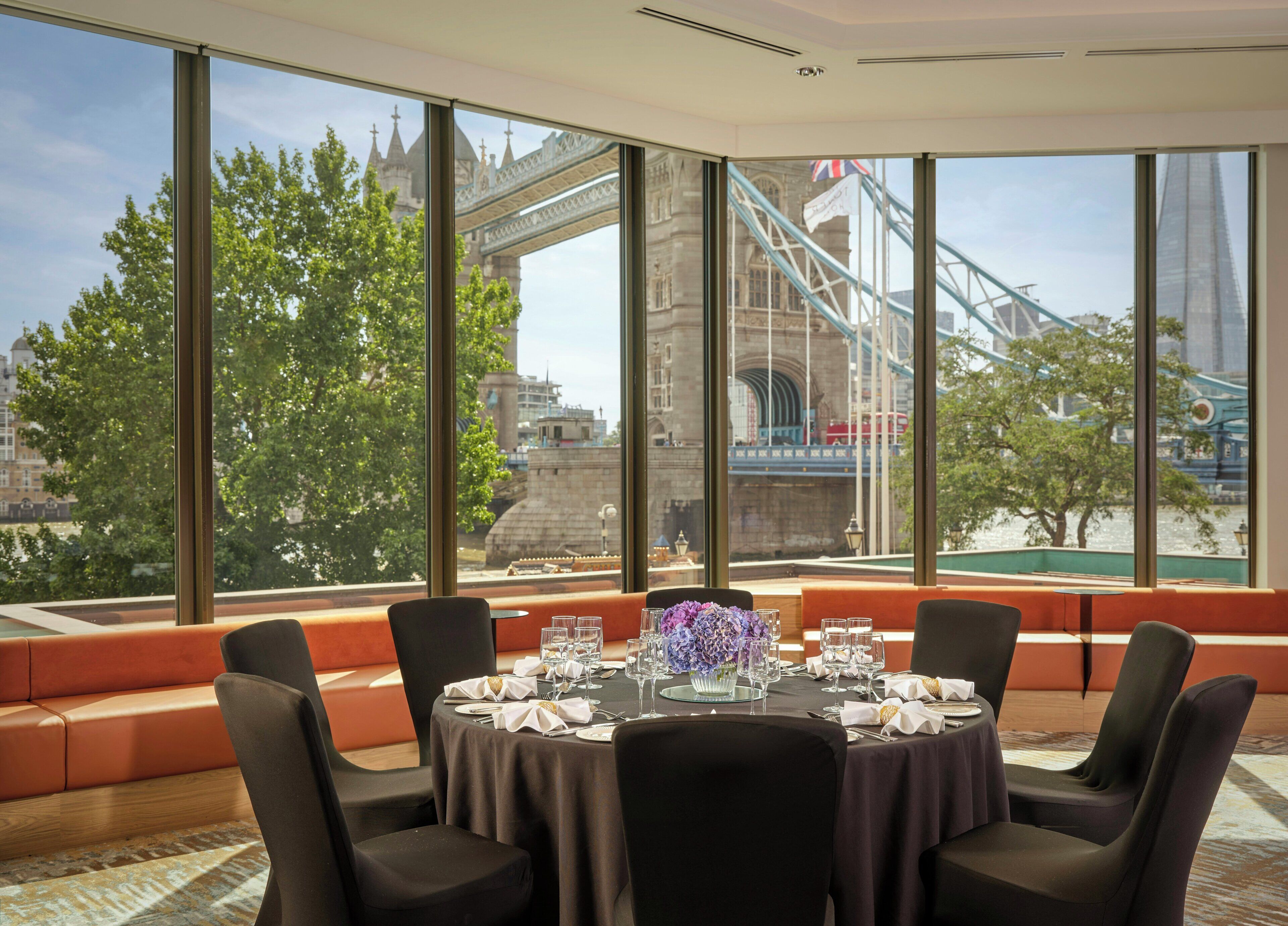Comedor con mesa para cuatro, ventanas amplias con vista a un puente y edificios, y zona de estar con cojines anaranjados.