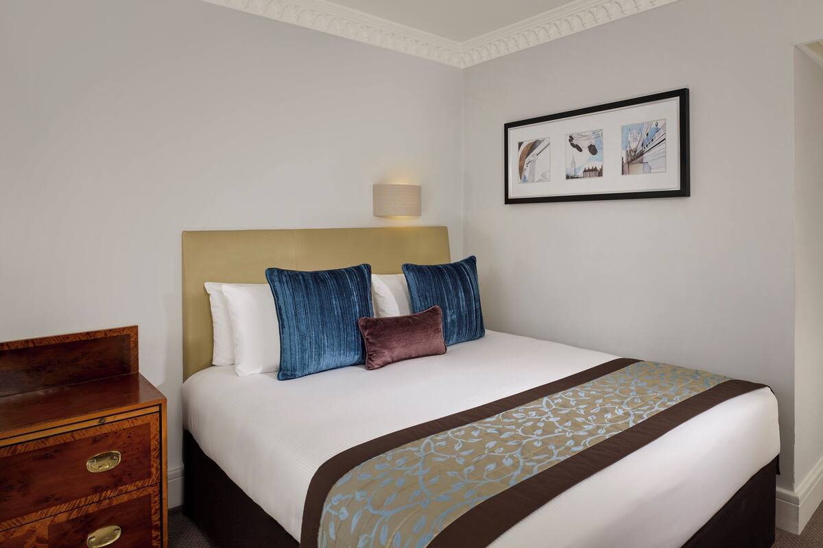 A neatly made bed with a patterned bedspread, a wooden dresser, and a framed picture on the wall.