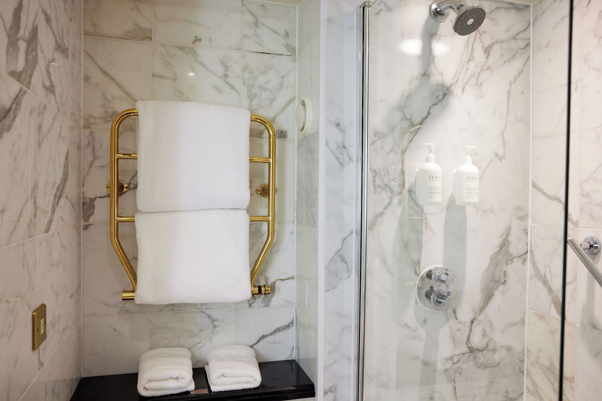 Une salle de bain aux murs de marbre, dotée d’une douche en verre, d’un porte-serviettes doré et d’une étagère noire où sont pliés les serviettes.