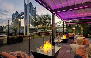Une terrasse extérieure dotée de tables avec foyer intégré, de sièges et offrant une vue sur un pont historique.