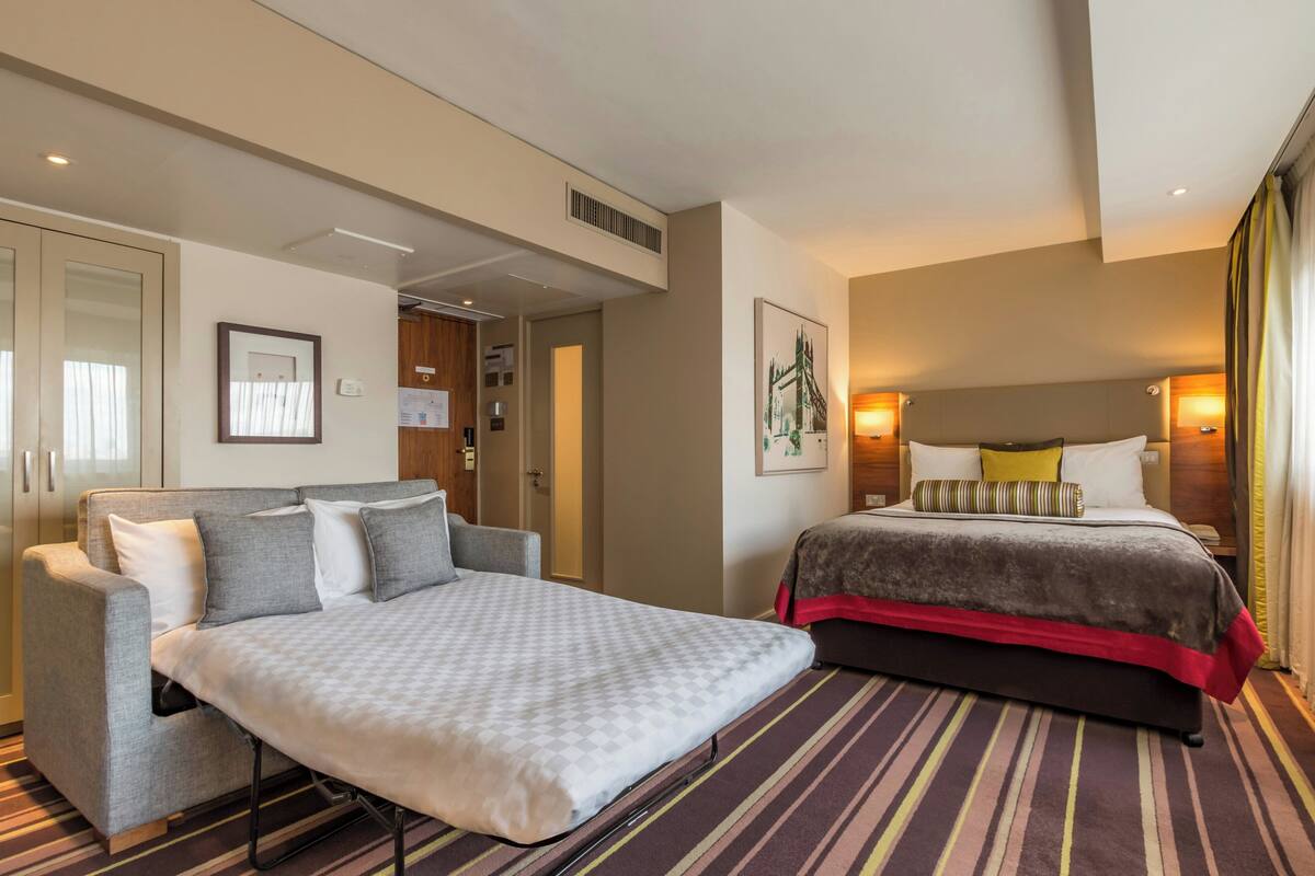 A hotel room with a bed, a sofa, a large window, and a door.