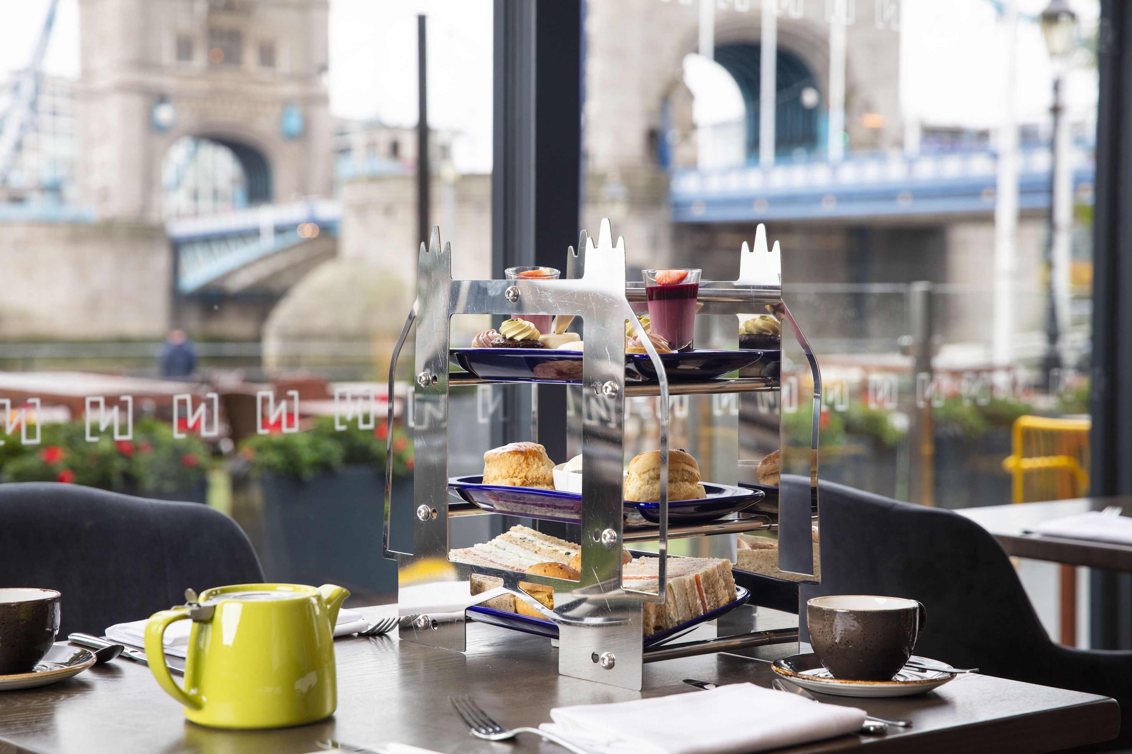 Eine gestufte Dessertauslage mit Sandwiches und Gebäck, ein gelber Teekessel und eine Tasse Kaffee auf einem Tisch mit Blick auf eine Brücke und Gebäude.