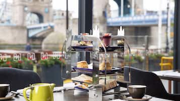 Un expositor escalonado con postres, sándwiches y pasteles, una tetera amarilla y una taza de café sobre una mesa con vistas a un puente y edificios.