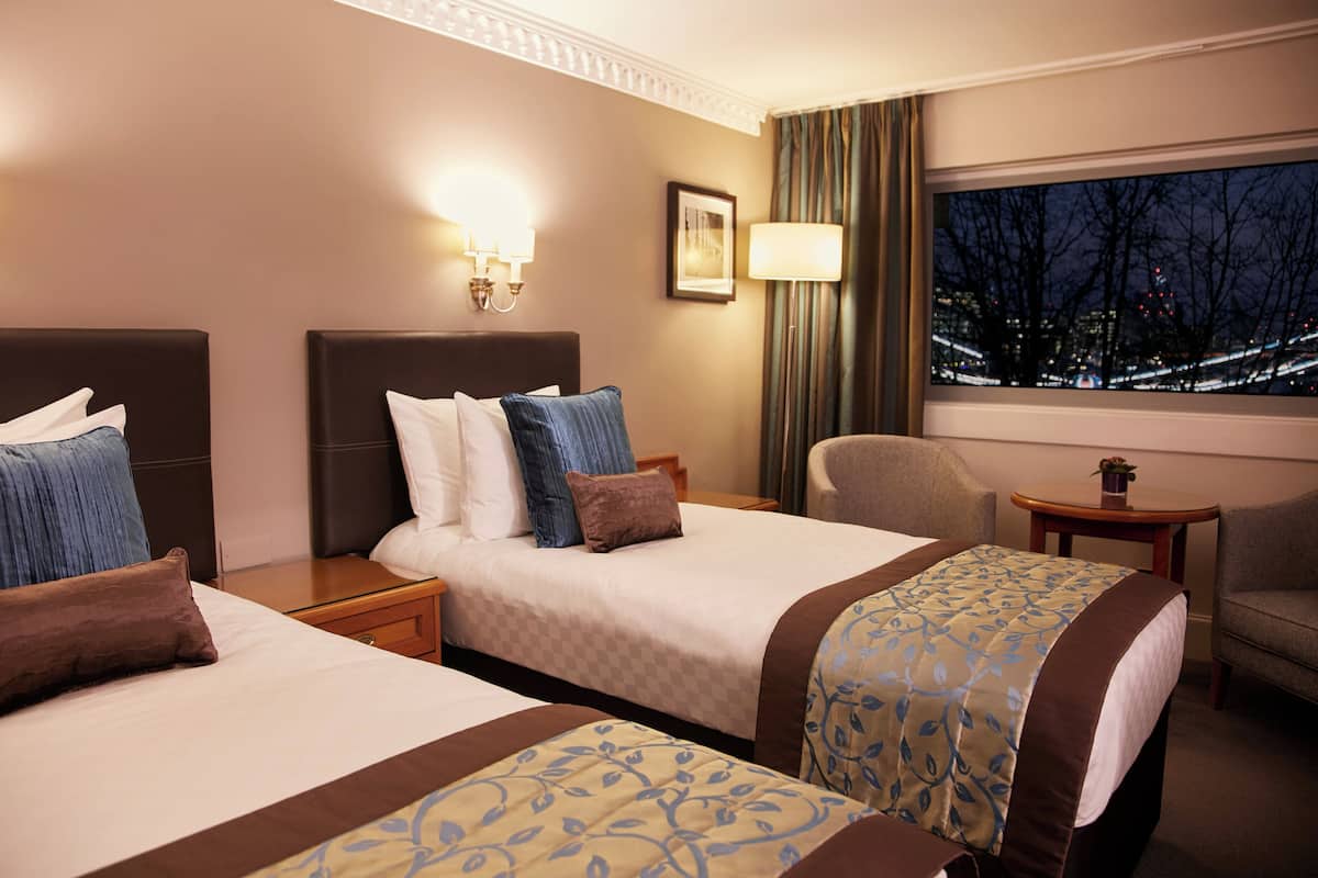 A hotel room with two beds, a sitting area, a window with a city view, and a lamp on the wall.