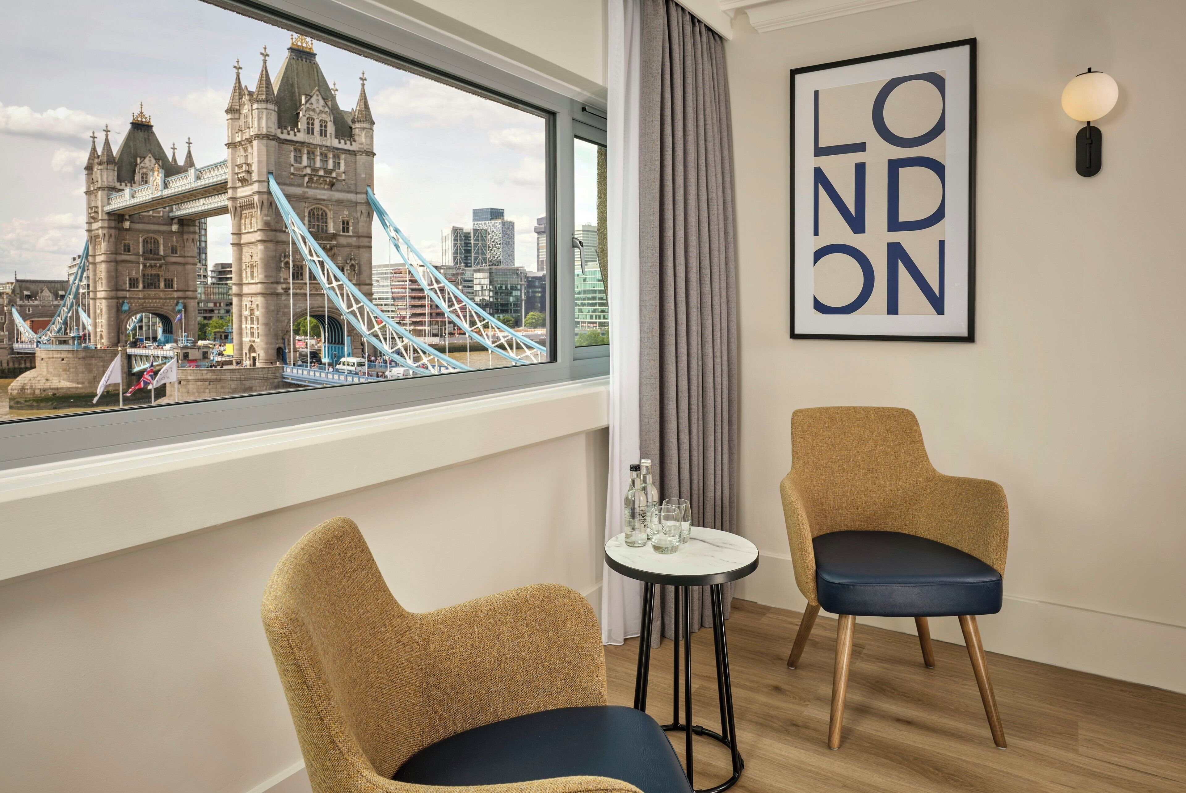 Una habitación con vistas al Tower Bridge, dos sillas, una mesita con vasos y botellas, y un cartel enmarcado con la palabra 'LONDON'.