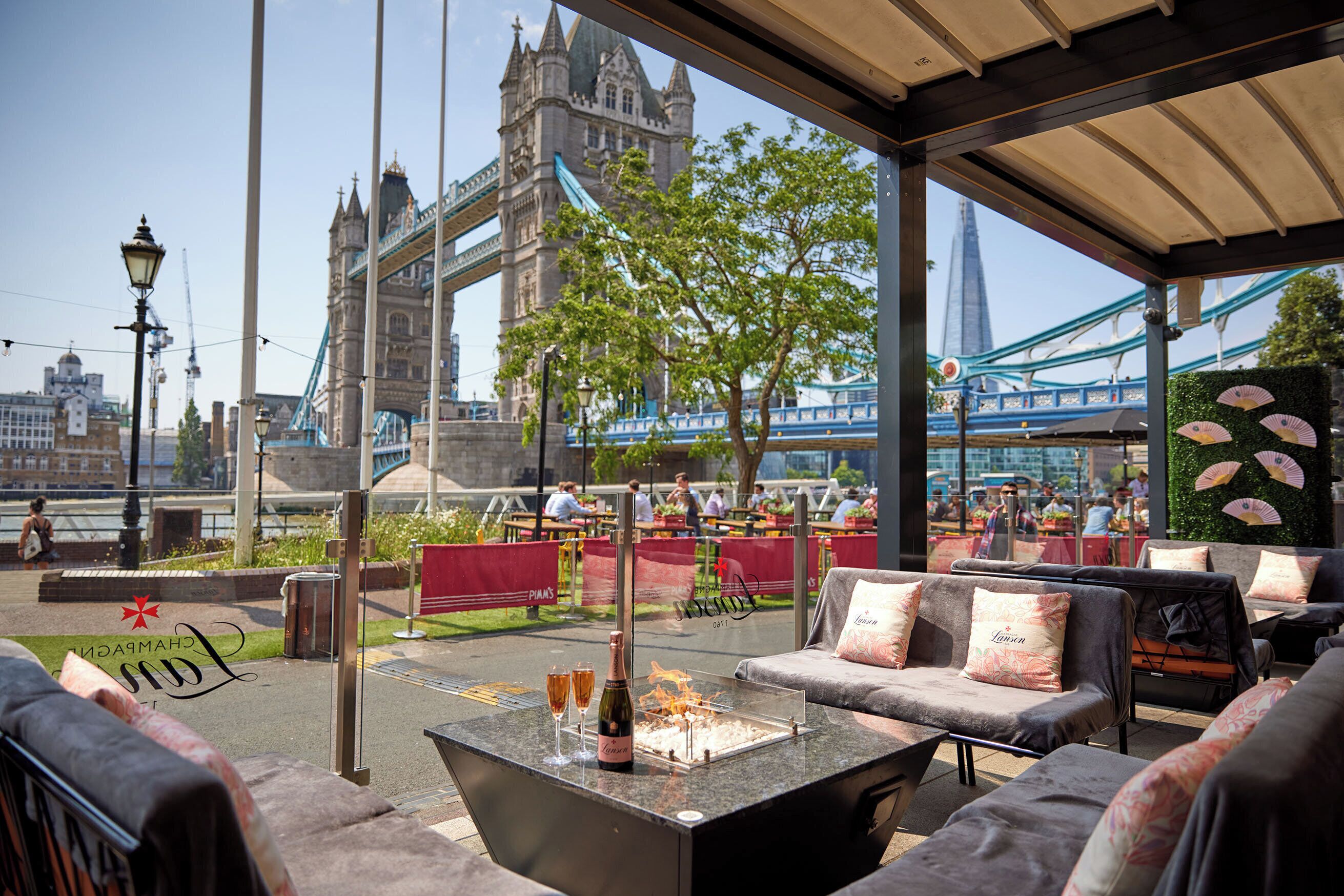 Un espace extérieur aménagé avec des sièges, offrant une vue sur le Tower Bridge, une bouteille de champagne et une bouteille de vin posées sur une table.