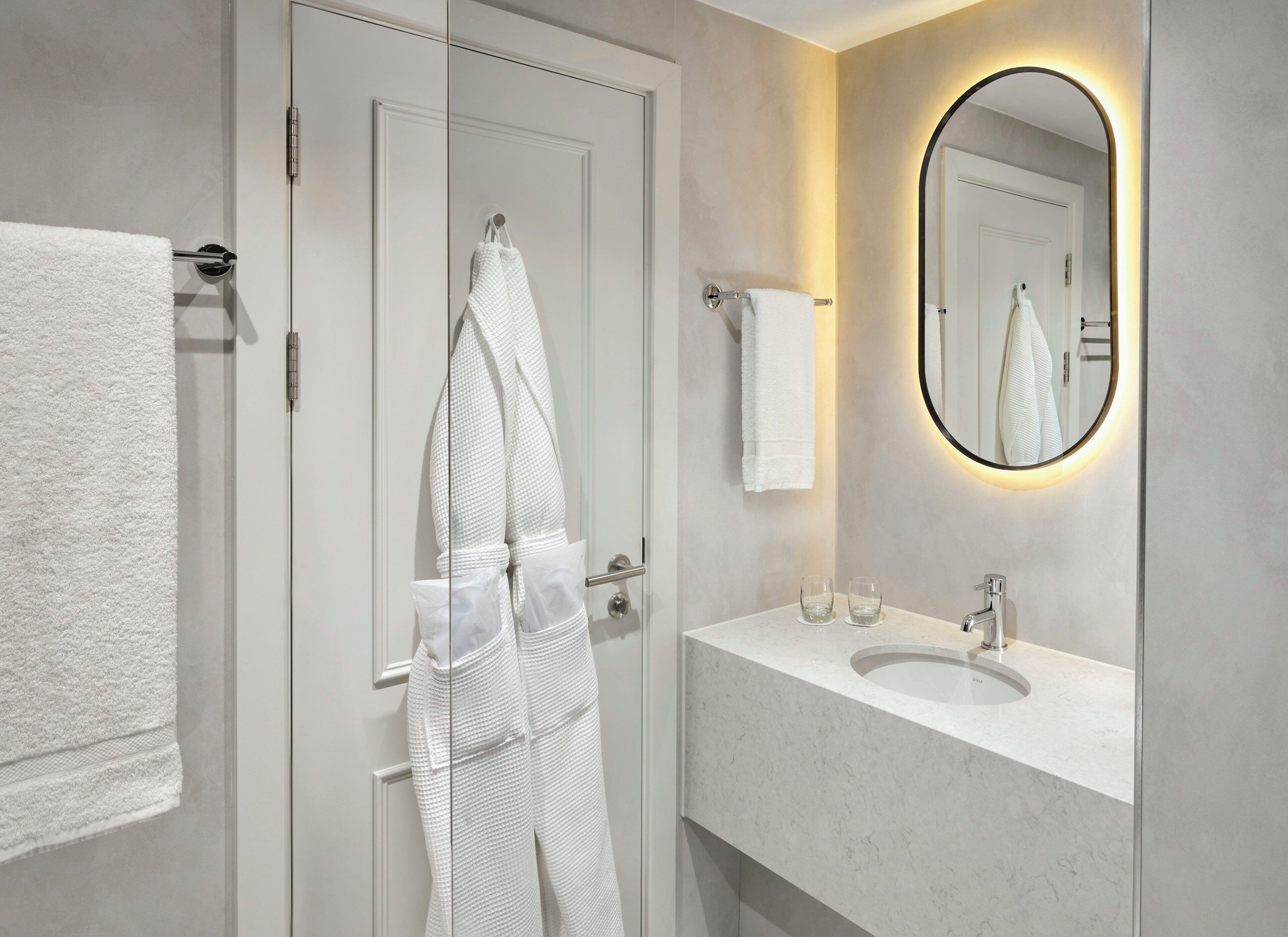 A modern bathroom with a white marble sink, a round mirror with a light halo, and a towel rack with white towels.