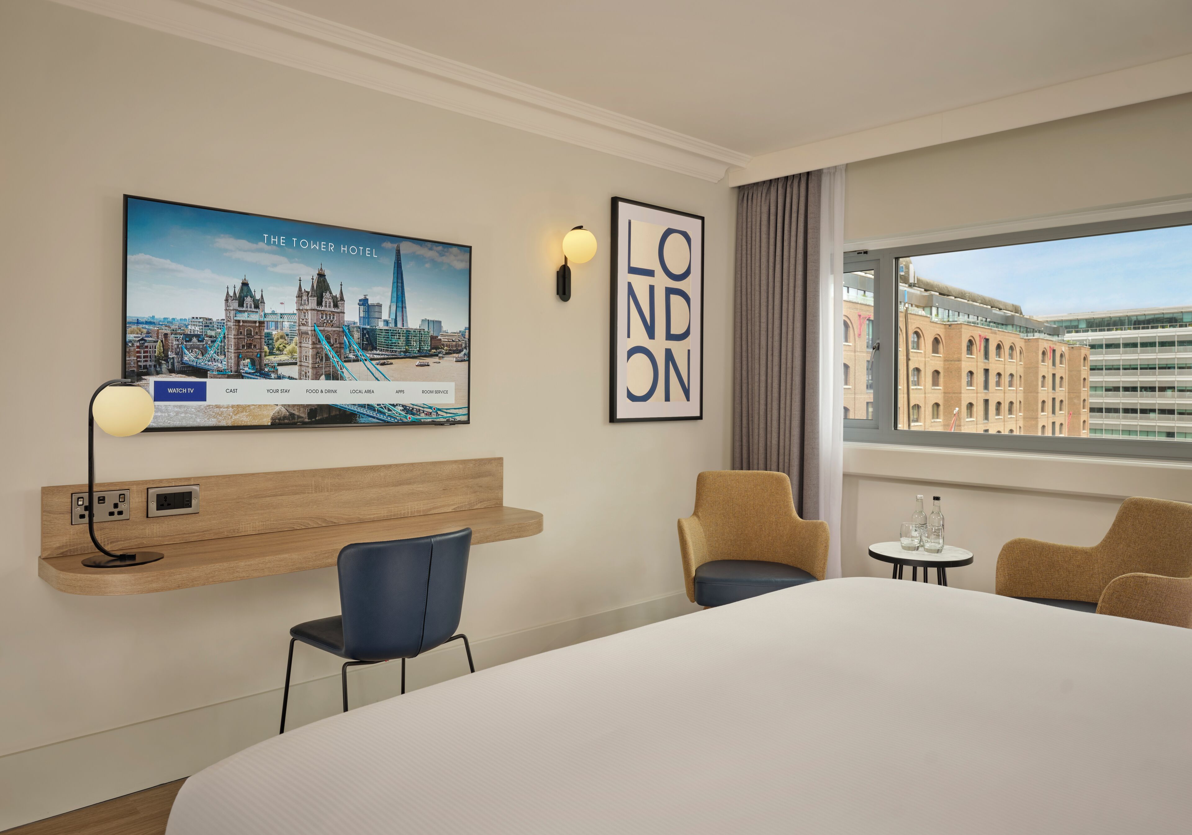 A hotel room with a large bed, a desk with a lamp, a TV displaying a cityscape, a framed sign with 'LONDON', and a window with a view of buildings.
