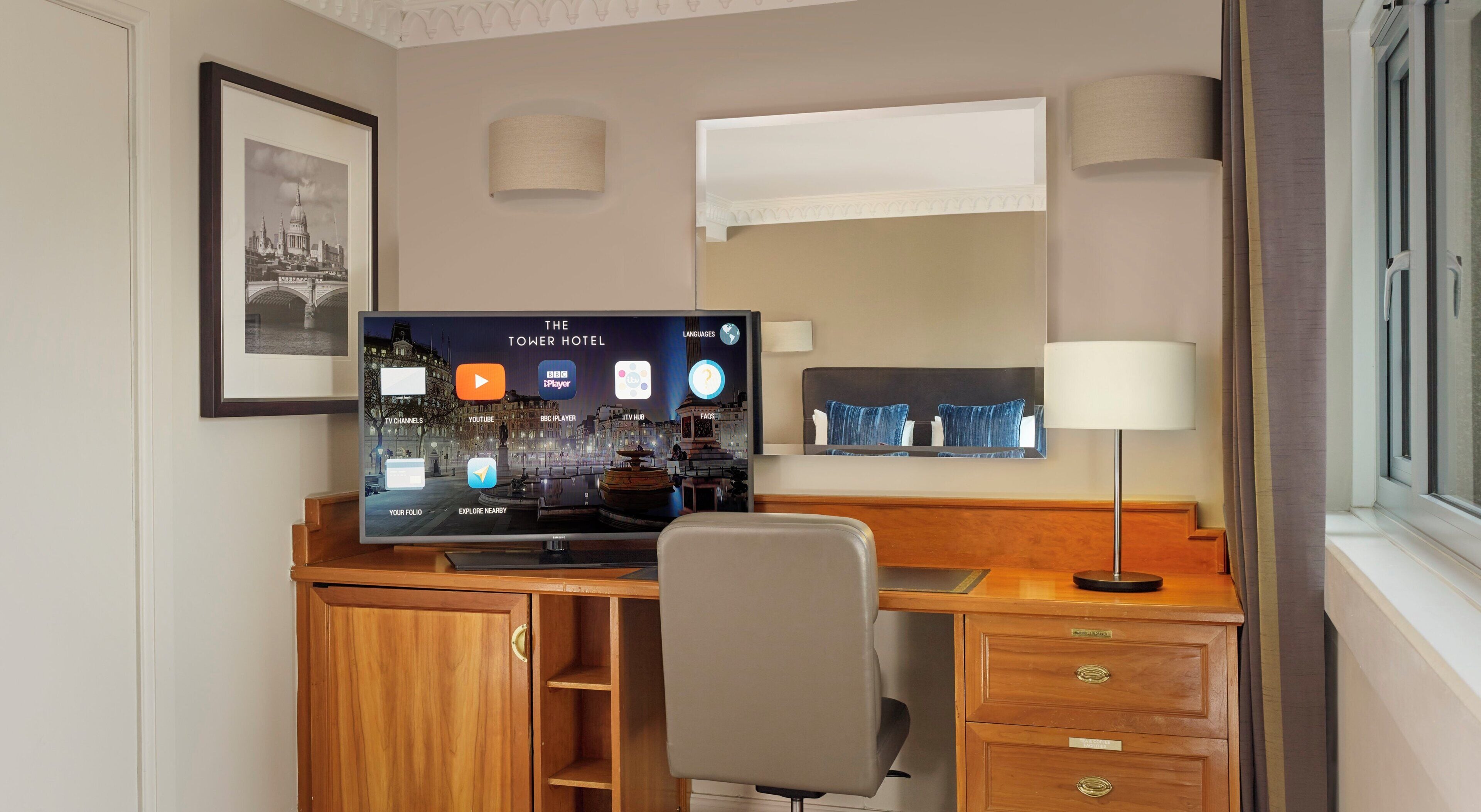 Une chambre d’hôtel avec un bureau en bois, un téléviseur à écran plat affichant une interface numérique, une chaise, une lampe et un tableau encadré au mur.