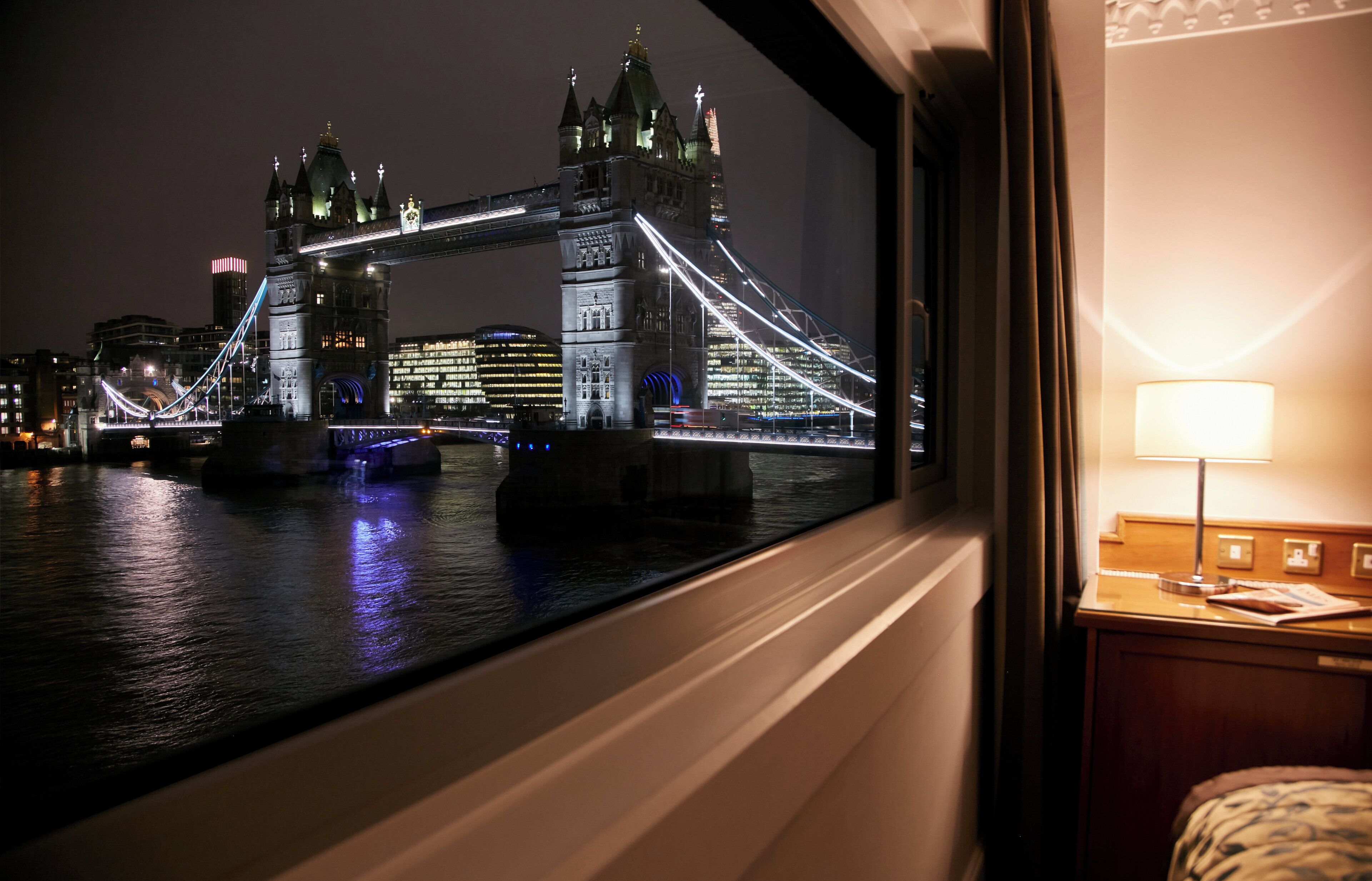 Vista del Tower Bridge por la noche desde una habitación con una lámpara en una mesita de noche.