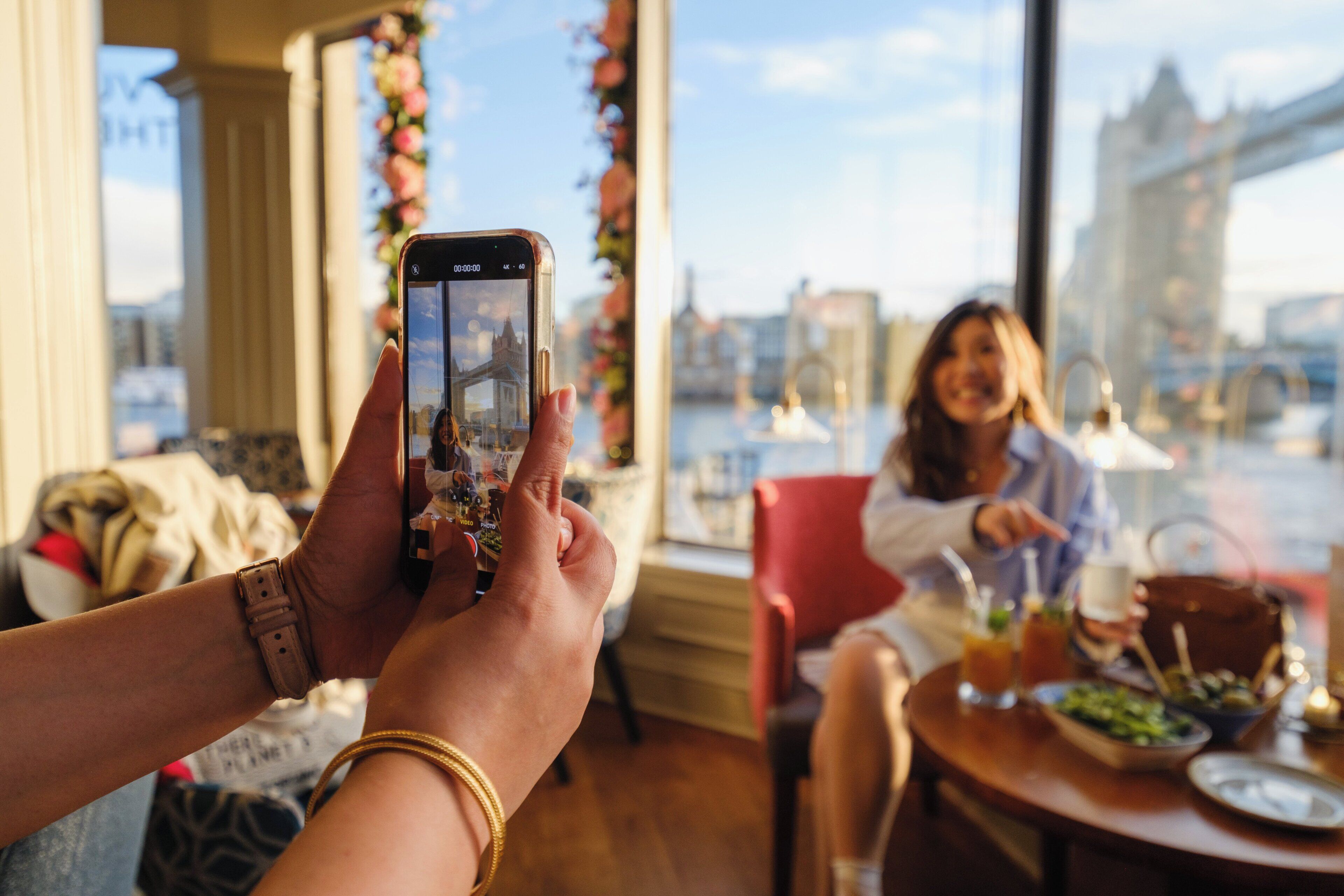 A person taking a photo with a smartphone, a dining table with food and drinks, and a view of a bridge and buildings through a window.