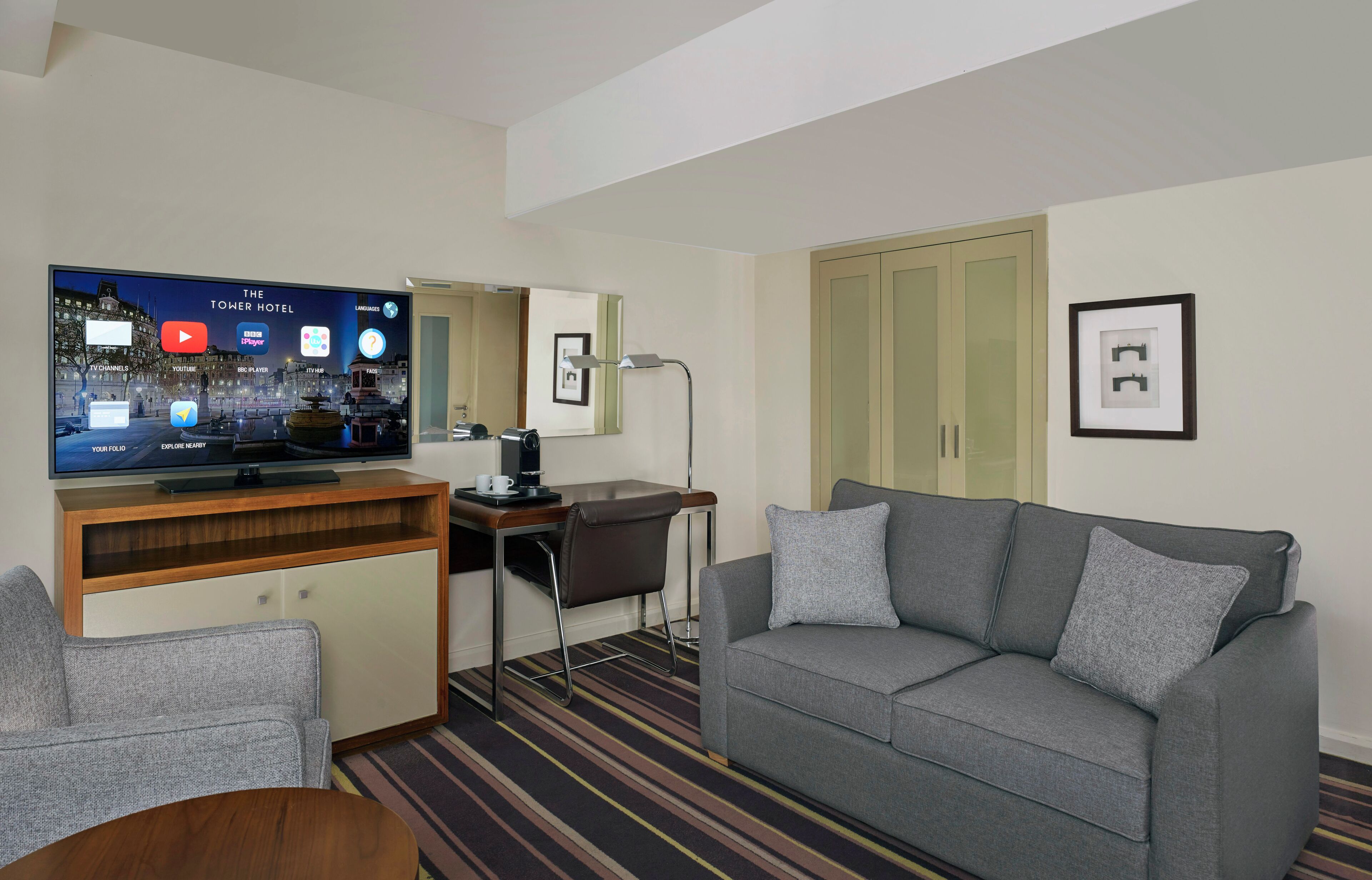 A hotel room with a TV displaying a hotel channel, a sofa, a desk with a chair, and a bed.