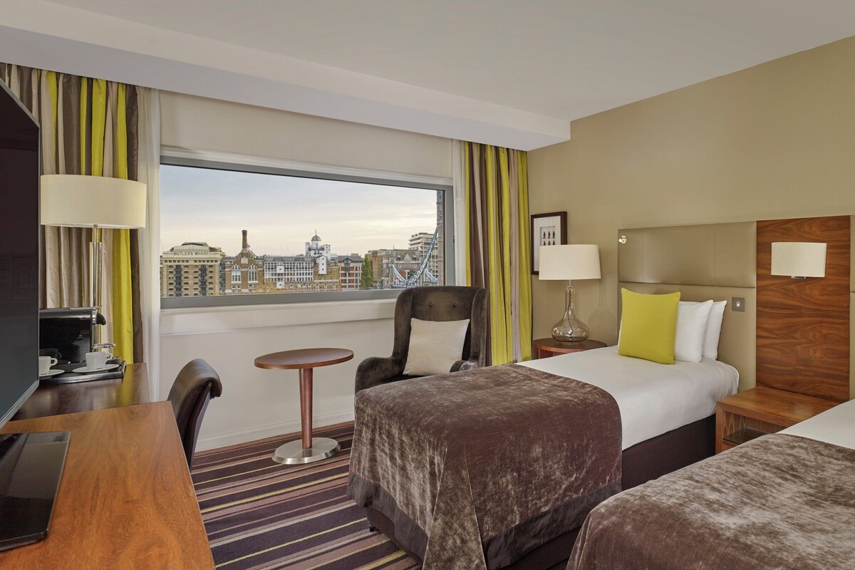 Quarto de hotel com cama, uma secretária com cafeteira, uma cadeira, uma mesa pequena e vista para a paisagem urbana.