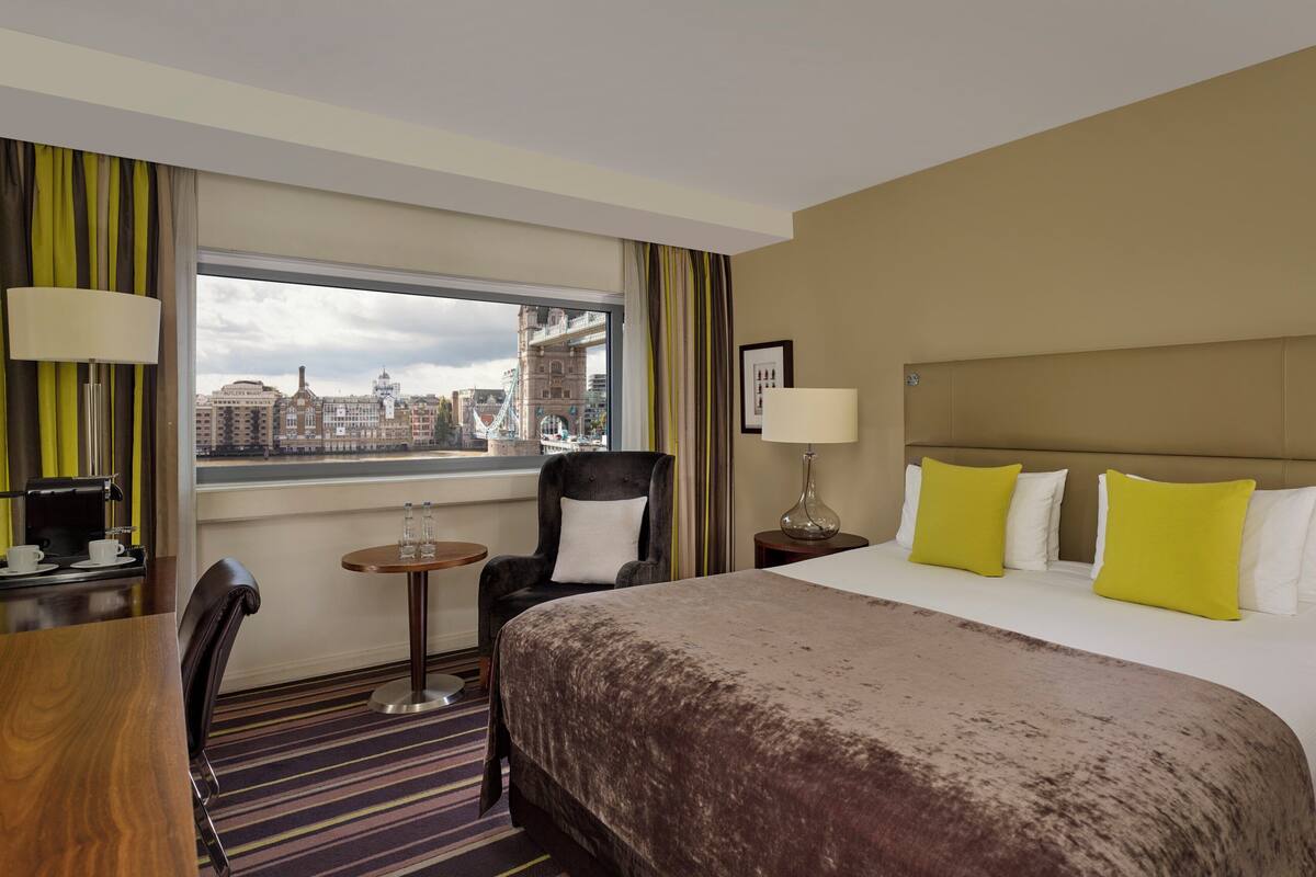 A hotel room with a bed, a chair, a table with a lamp, and a view of a cityscape.