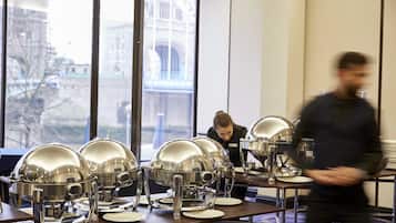 Ein Buffet mit mehreren Servierstationen, eine Person im Hintergrund und Blick auf ein Gebäude durch große Fenster.