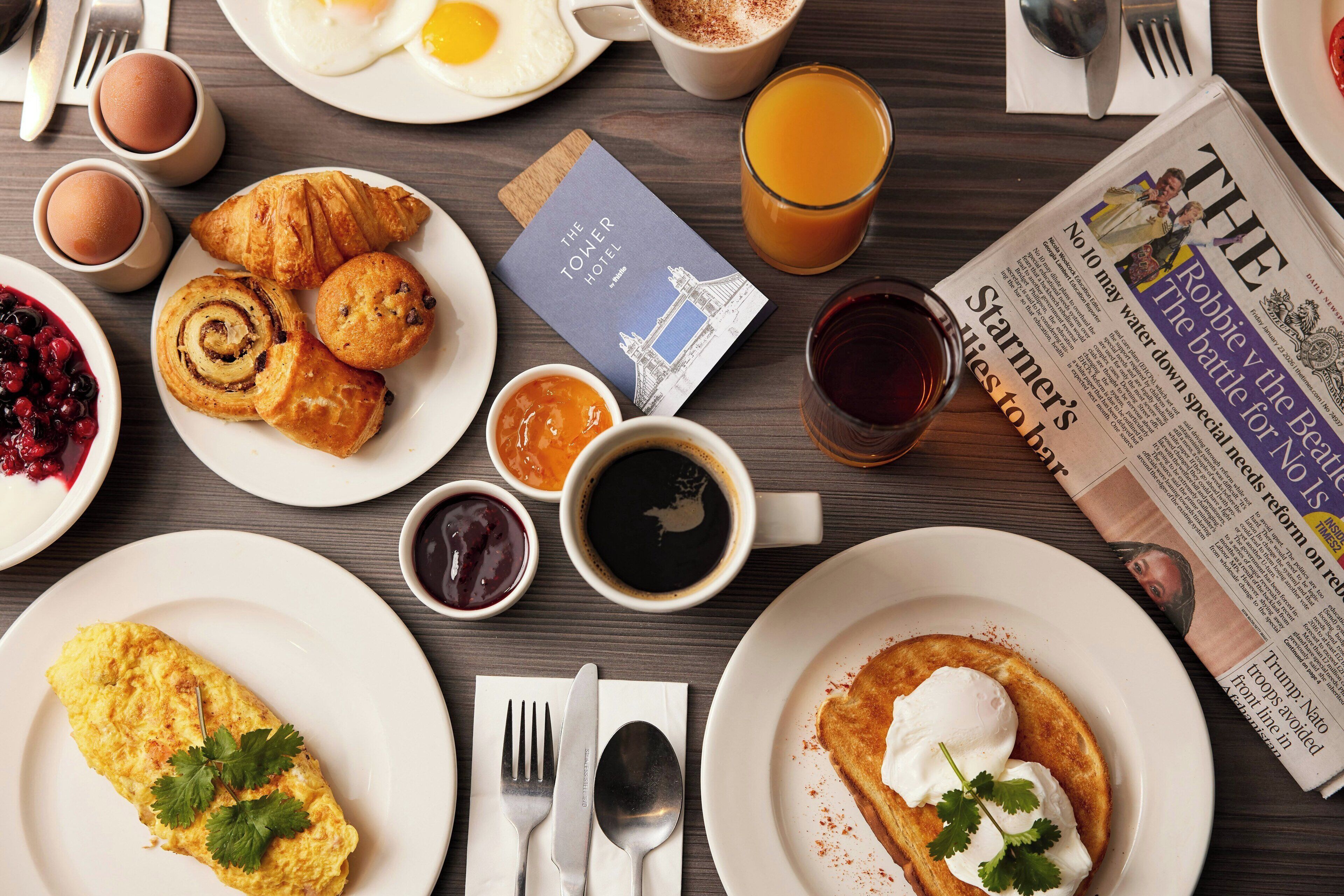 Mesa posta para o café da manhã, com diversos alimentos, um jornal e um livro.