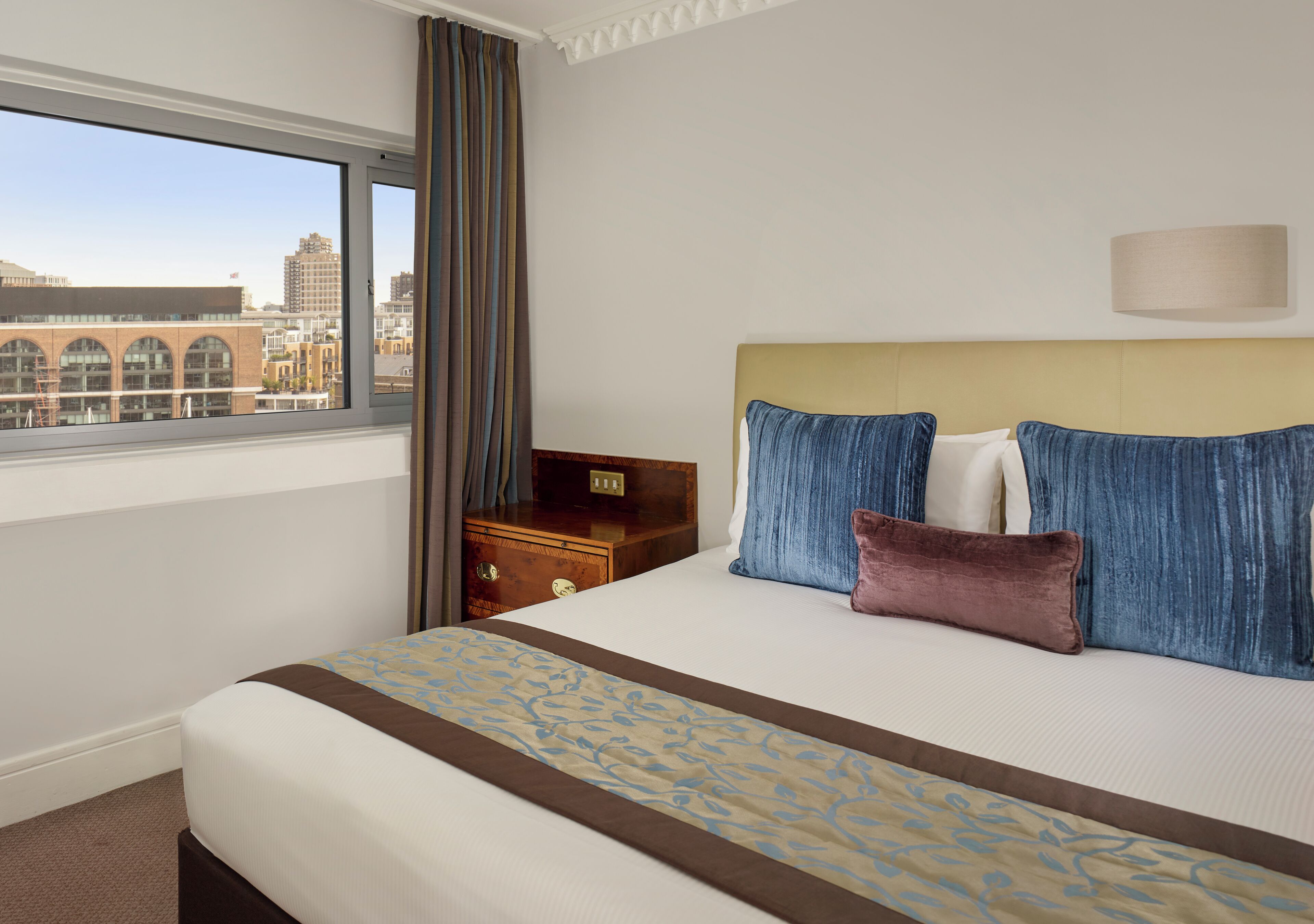 A hotel room with a large window, a bed with patterned bedding, a wooden nightstand, and a city view.