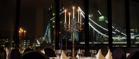 A table set for dining with a candelabra centerpiece, wine glasses, and a view of a bridge at night.