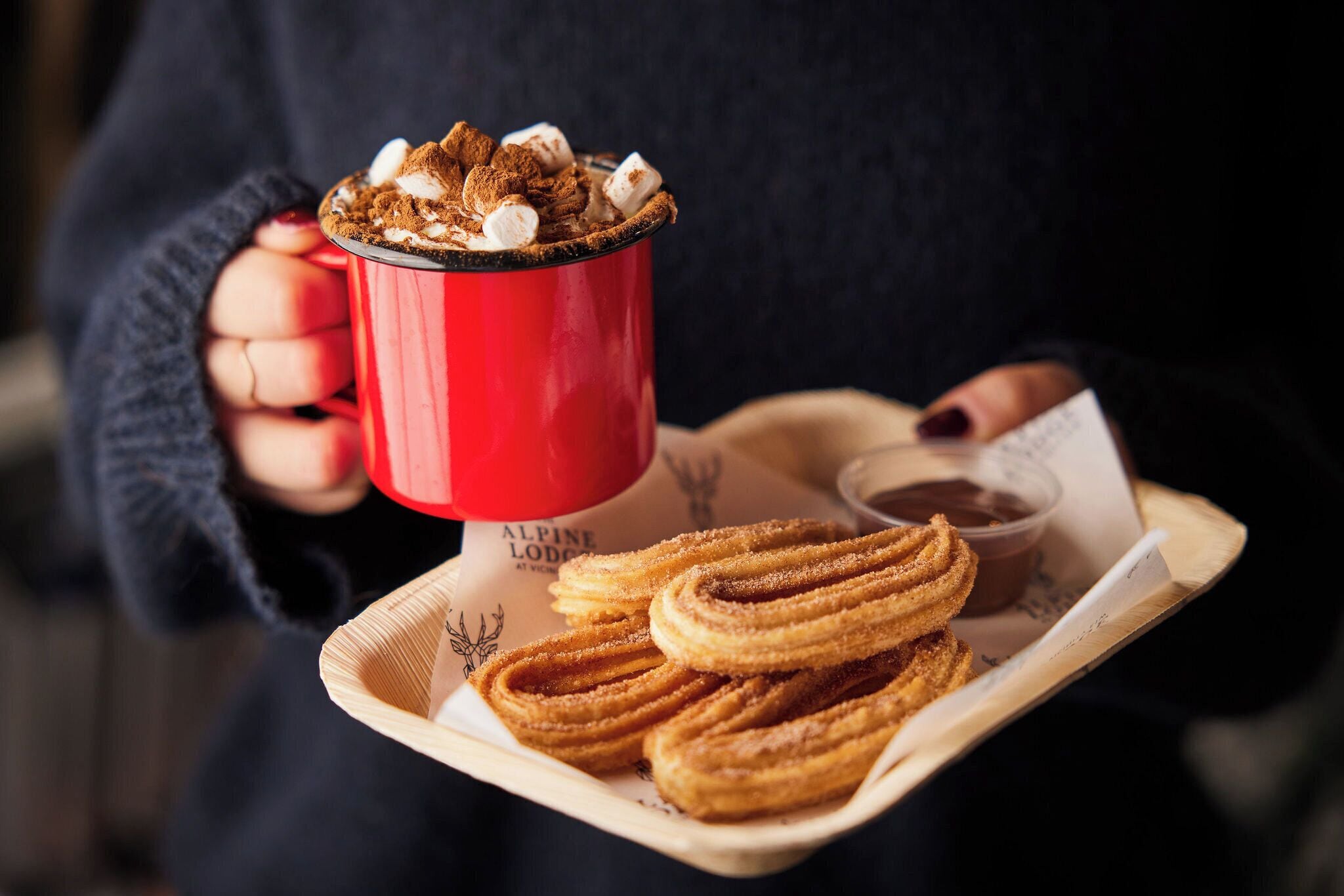 A person holding a red mug with a hot beverage and churros on a tray.