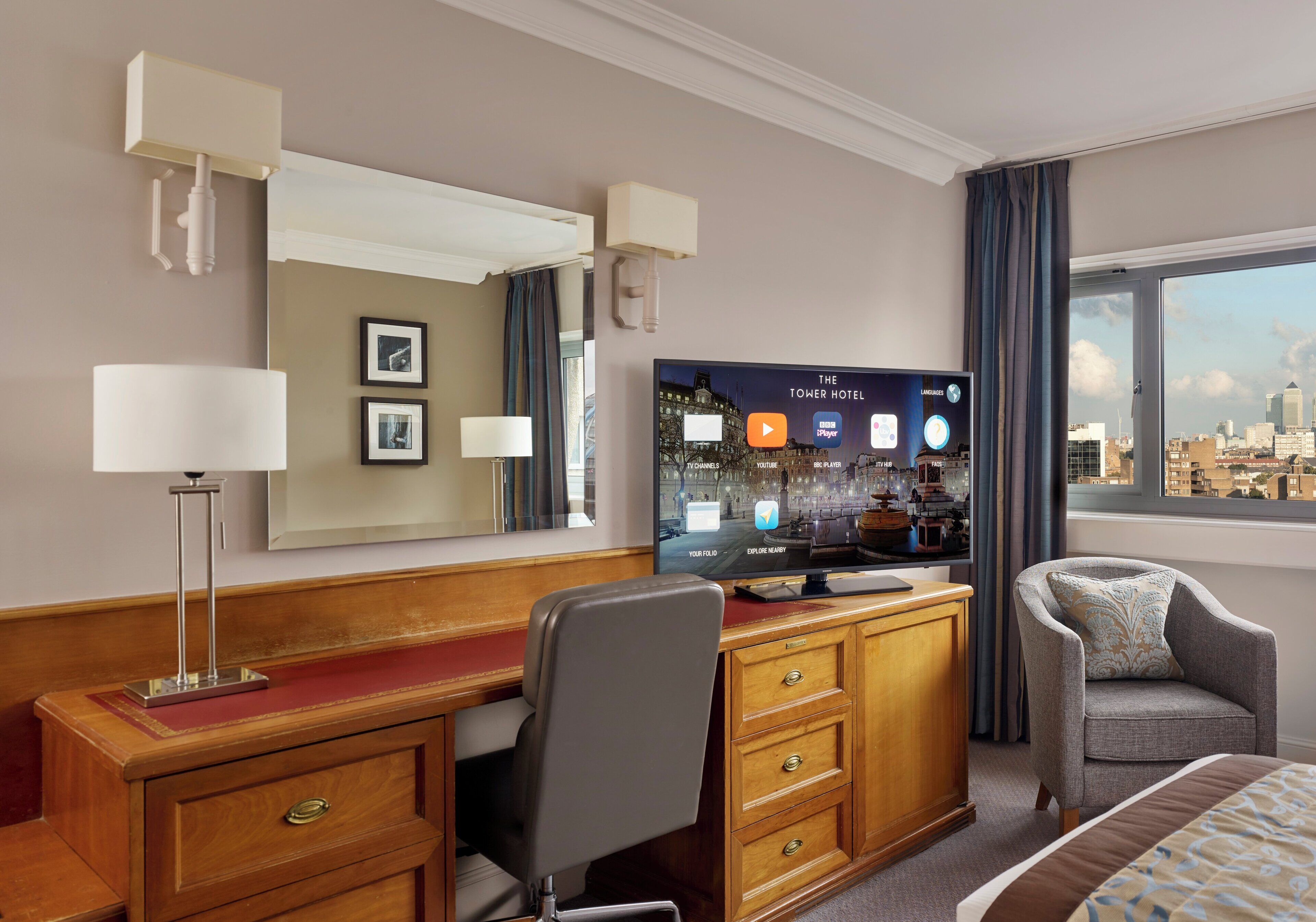 A hotel room with a TV, a desk, a chair, a bed, a large window, and a city view.