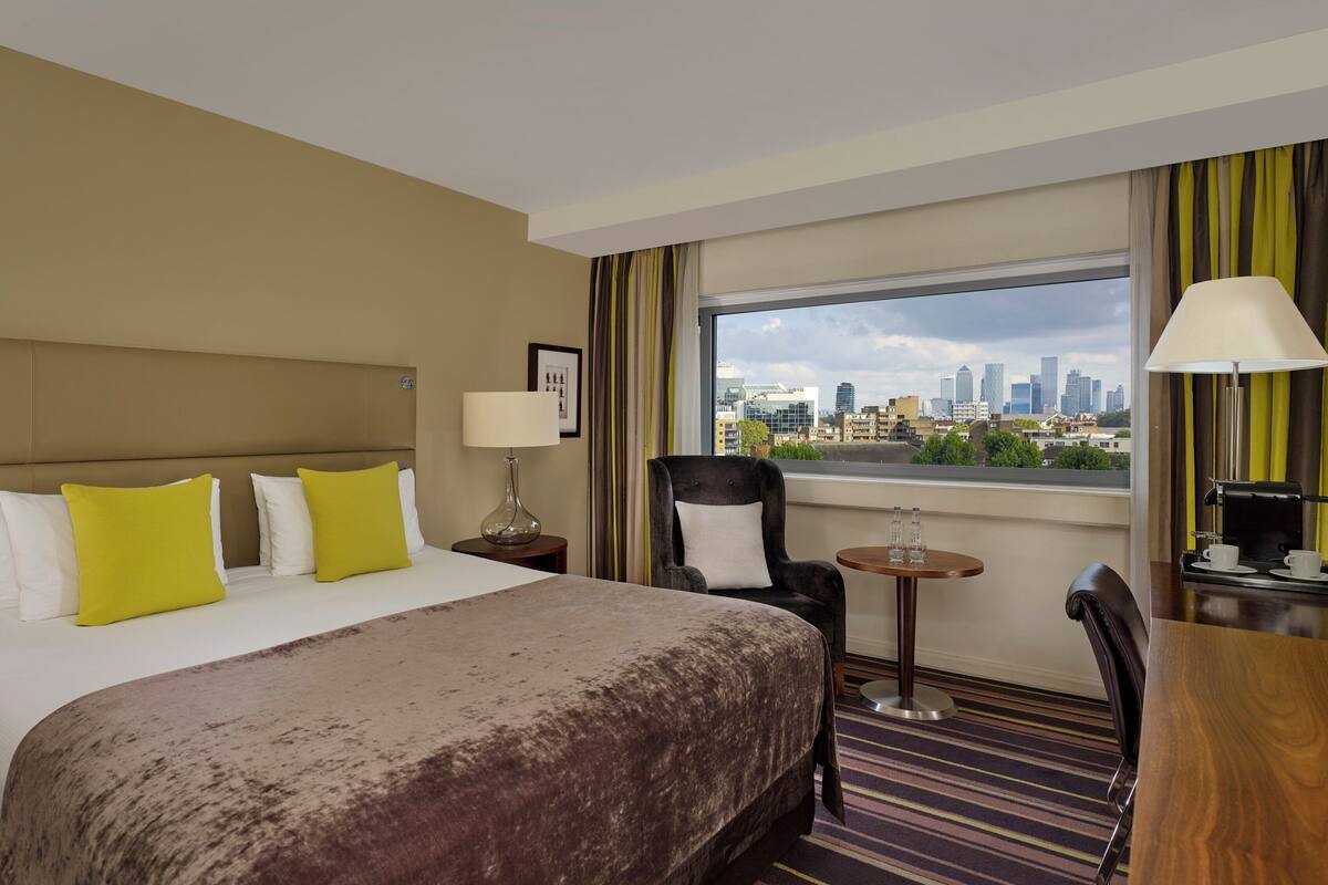 A hotel room with a bed, a chair, a table, a lamp, and a city view.