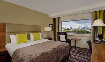 A hotel room with a bed, a chair, a table, a lamp, and a city view.