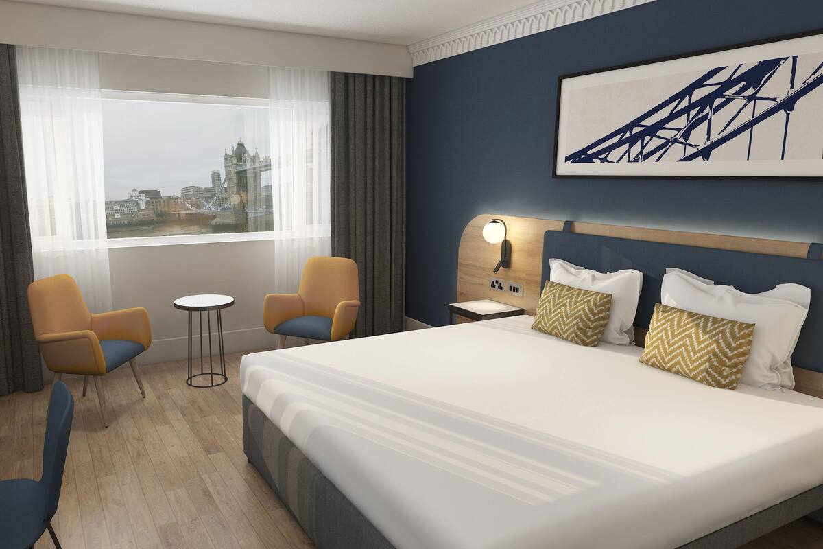 A hotel room with a large bed, two armchairs, a small table, a nightstand with a lamp, and a view of a bridge and buildings.
