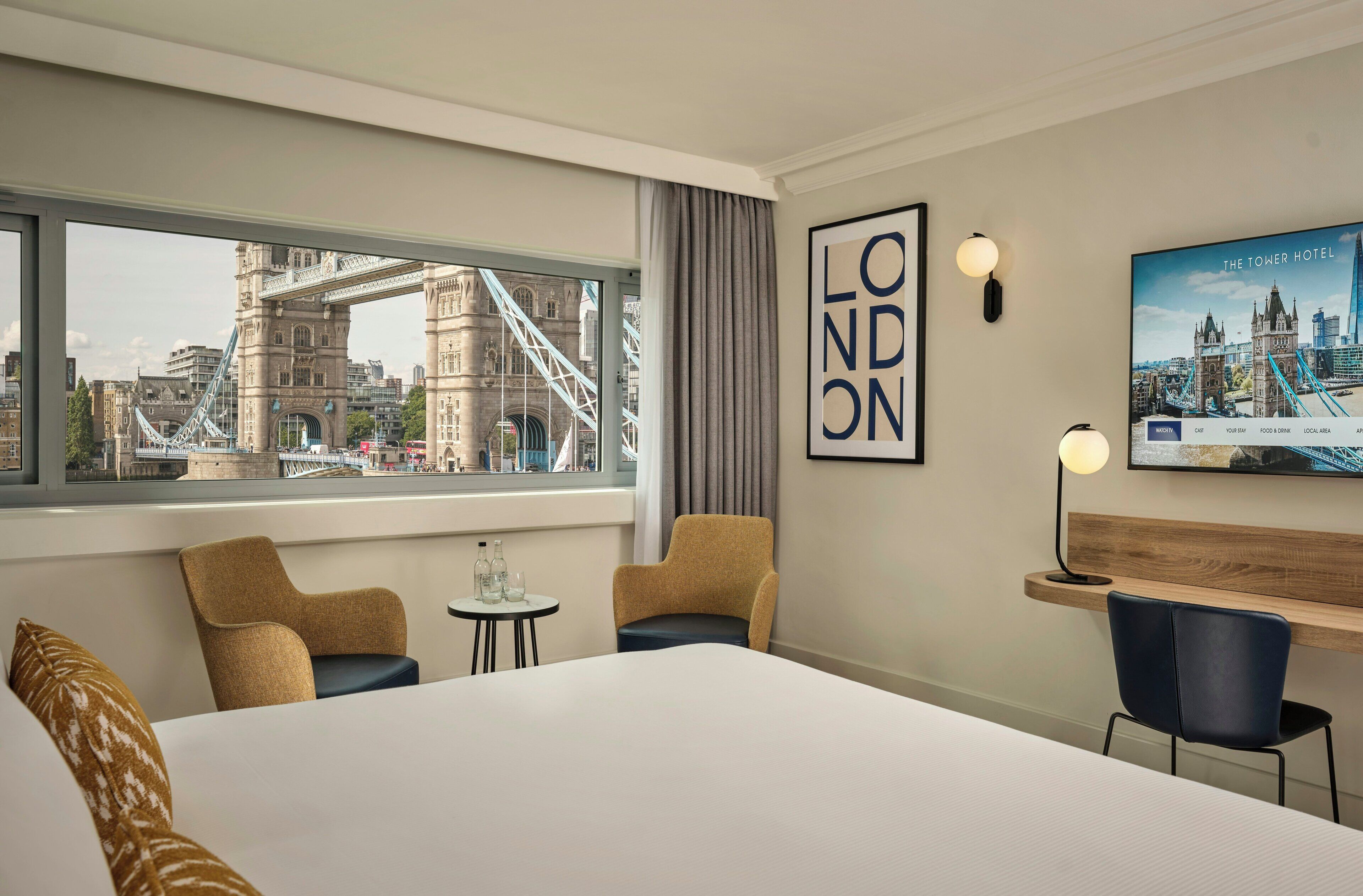 Habitación de hotel con una cama grande, dos sillones, una mesita con botellas de agua, un escritorio con una silla, una televisión mostrando un paisaje urbano y vistas de un puente y edificios.