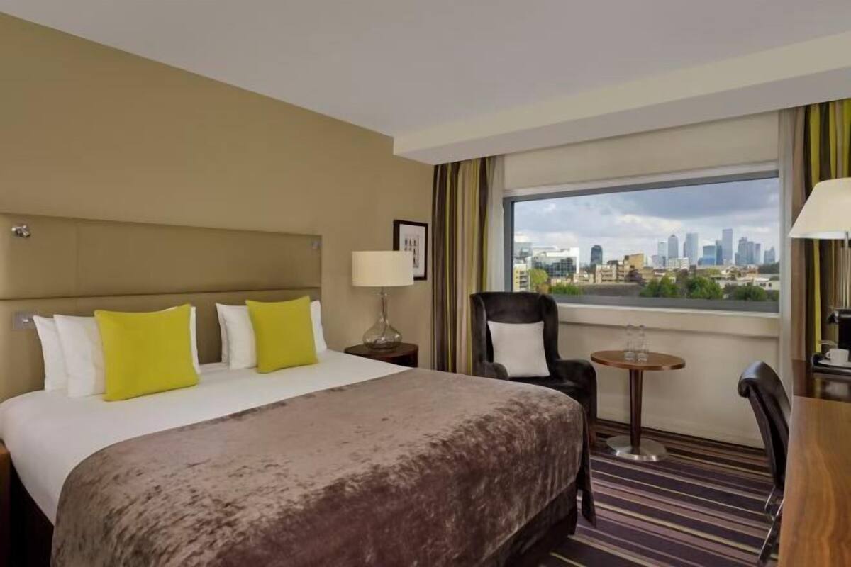 A hotel room with a large bed, a chair, a small table, and a view of a cityscape.