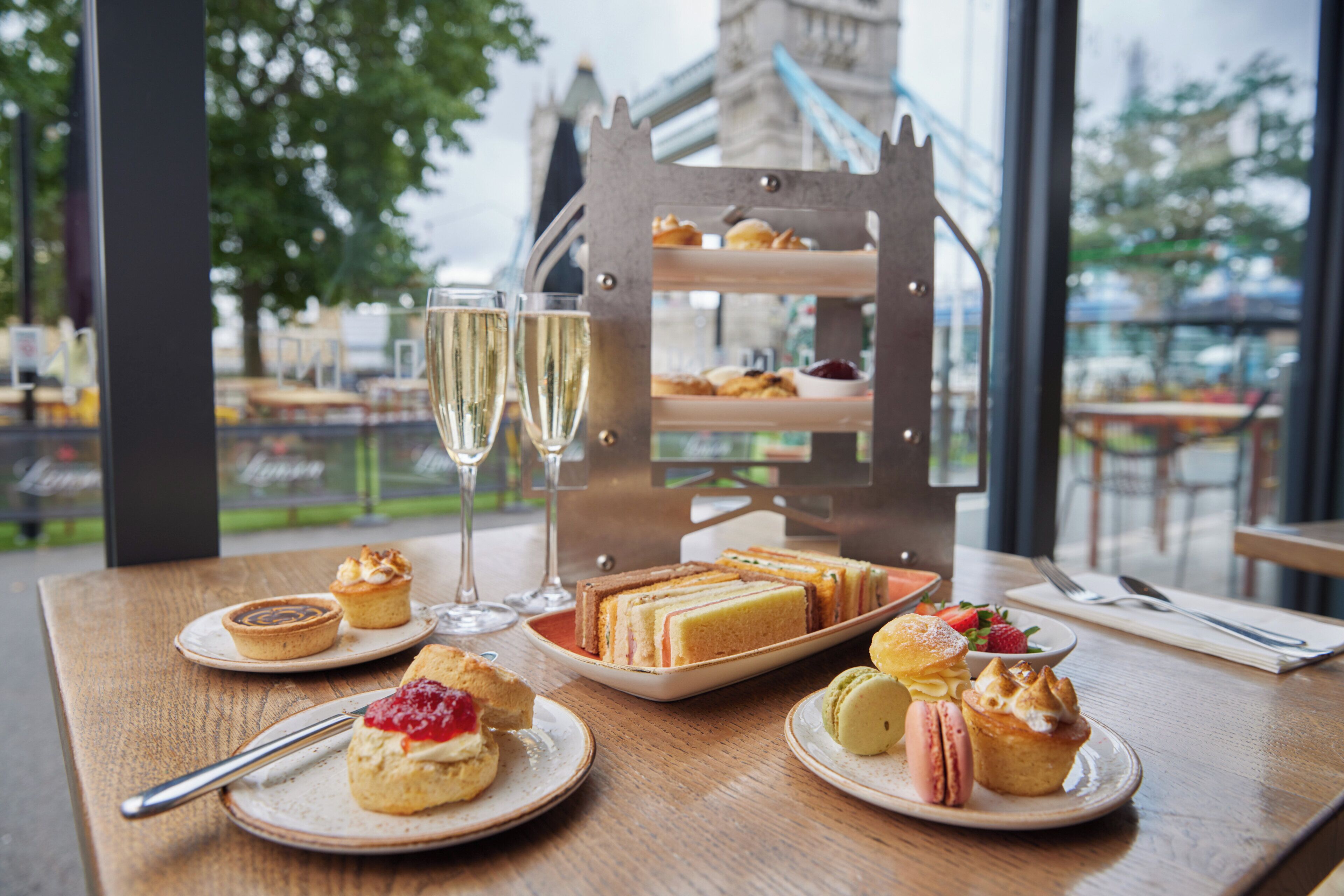 A table with various pastries and a tiered dessert stand, a glass of champagne, and a view of a bridge in the background.