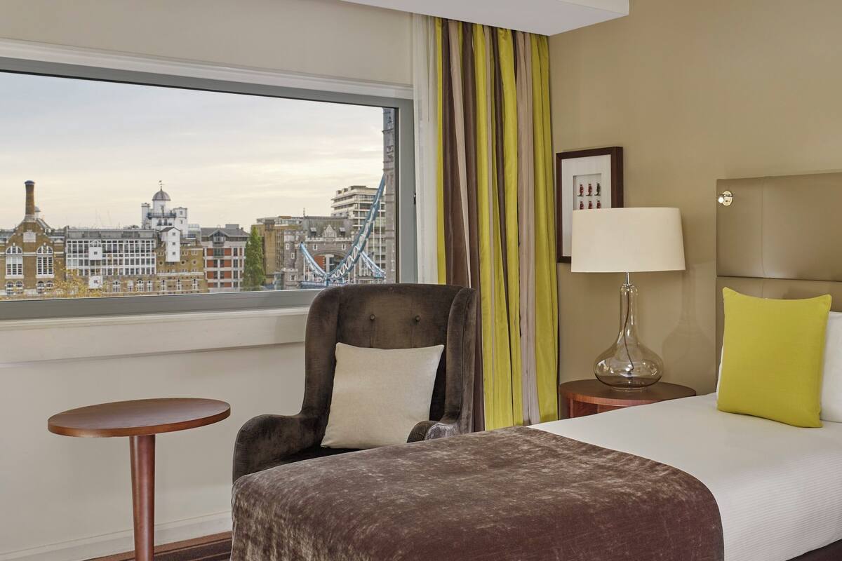 A hotel room with a bed, a chair, a small table, a lamp, and a view of a cityscape with a bridge.