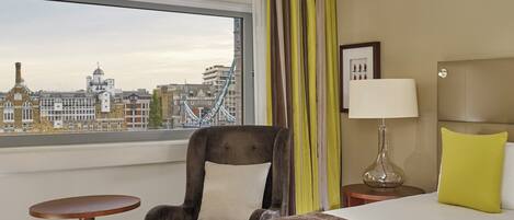 A hotel room with a bed, a chair, a small table, a lamp, and a view of a cityscape with a bridge.