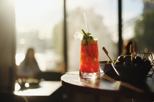 A glass of red beverage with a straw and mint garnish on a wooden table, with a bowl of olives and a metal rack in the background.