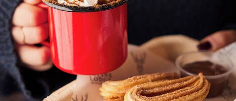 A person holding a red mug with a hot beverage, a plate with churros, and a small bowl of sauce.