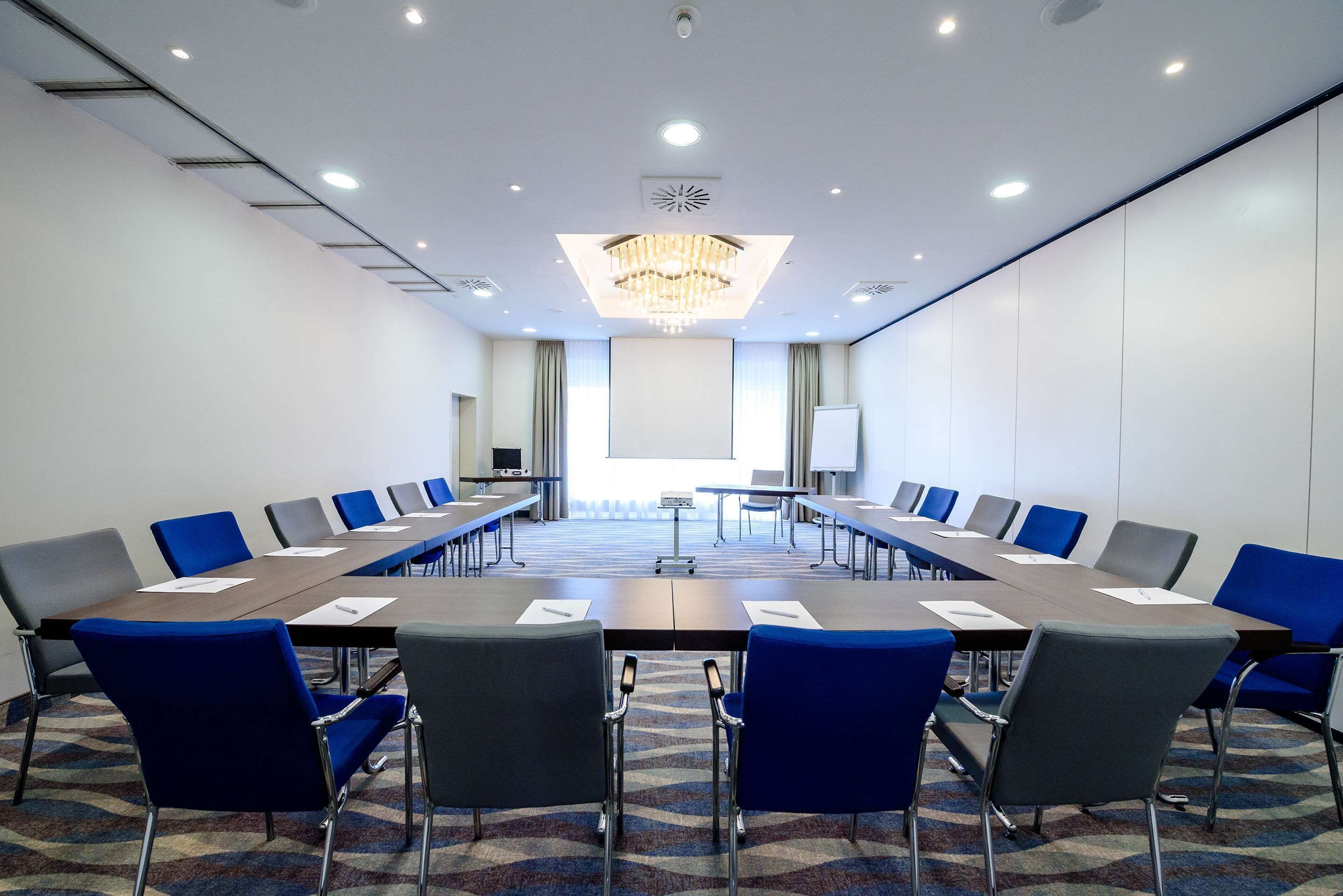 Une salle de conférence avec une longue table, des chaises bleues et grises, un lustre et un écran de projection.