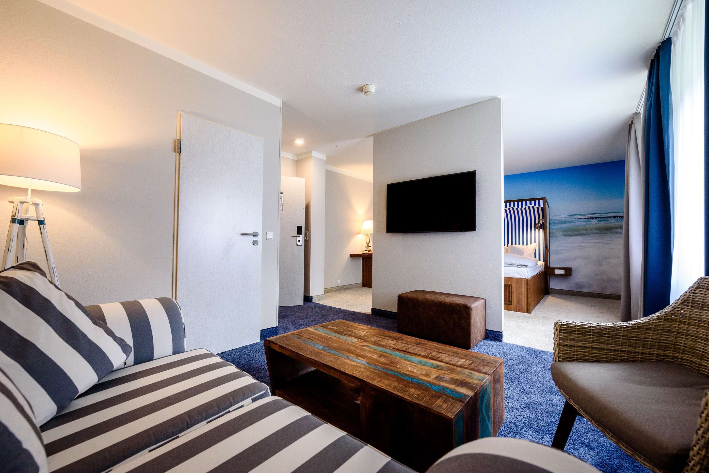 A hotel room with a striped sofa, a wooden coffee table, a wicker chair, a flat-screen TV mounted on the wall, and a view of the sea through the window.