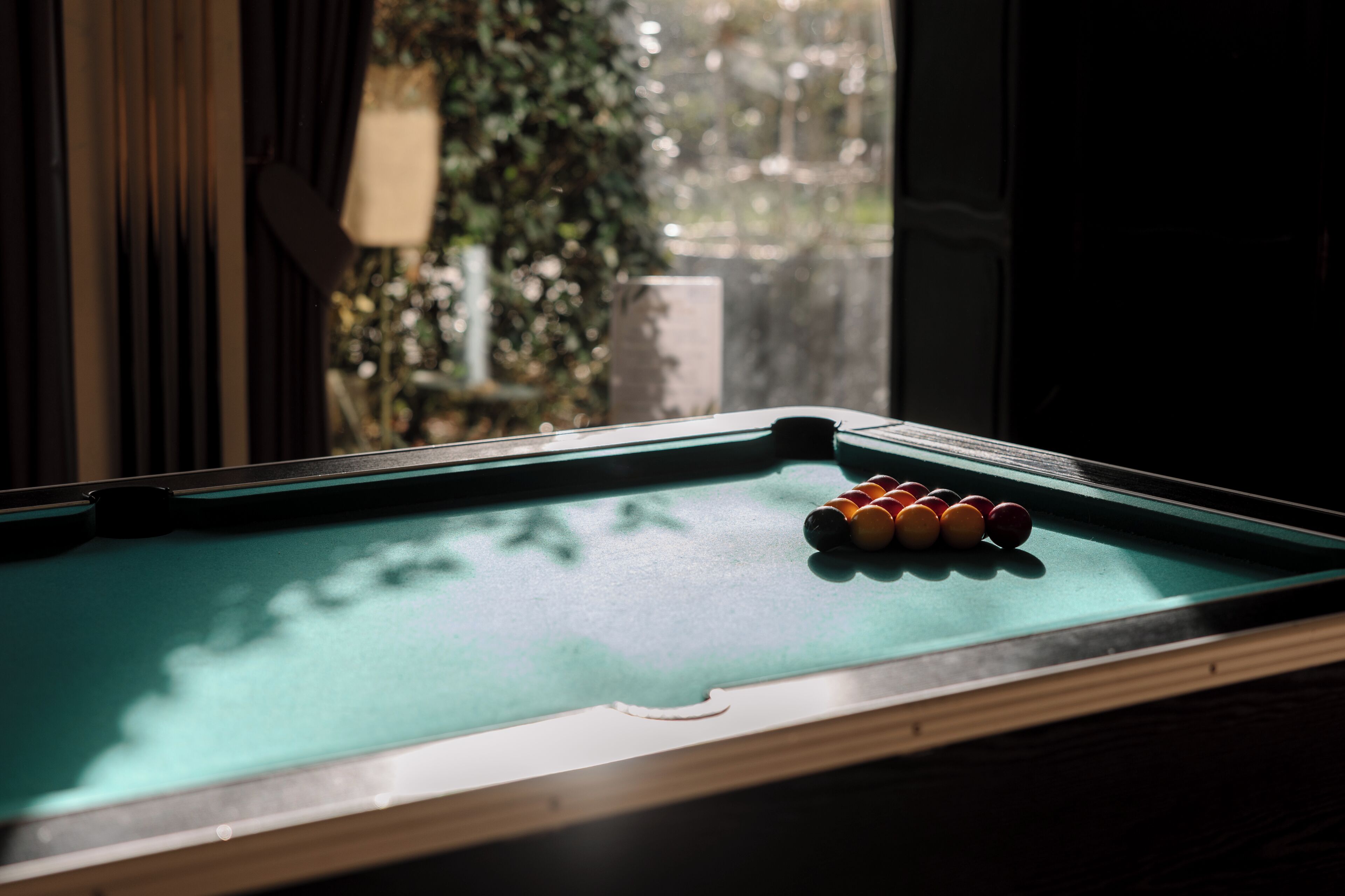 Une table de billard avec les billes disposées, une fenêtre avec des stores et une vue sur la verdure à l’extérieur.