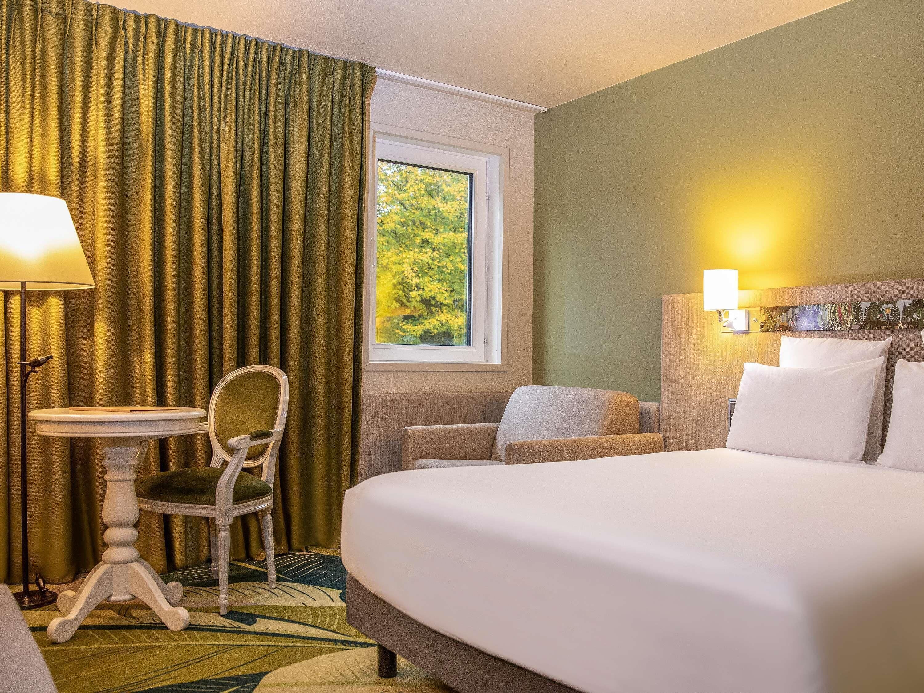 Une chambre d’hôtel avec un lit, une chaise, une petite table, une fenêtre donnant sur des arbres et une lampe.