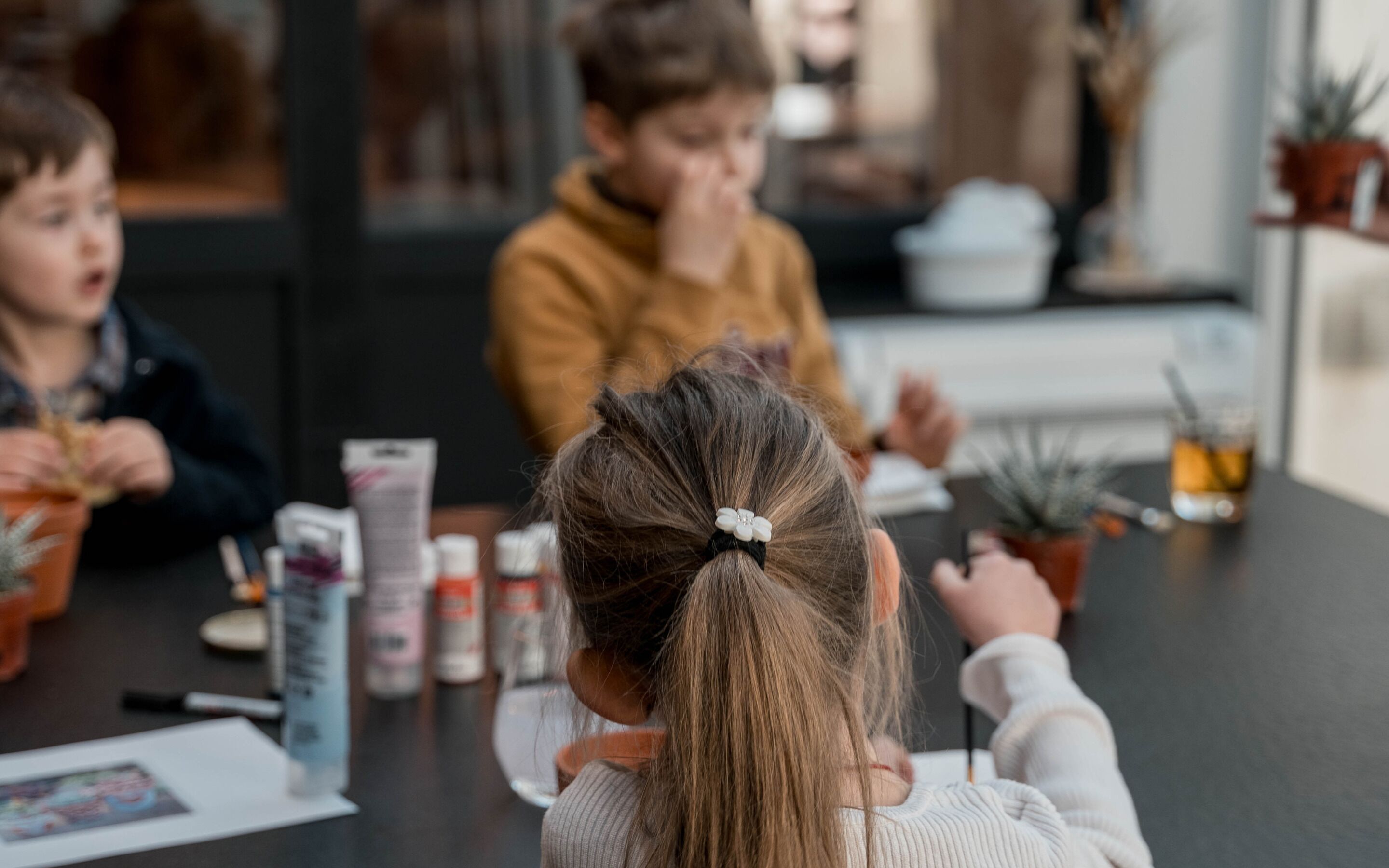 A child with a ponytail sitting at a table with a potted plant, a notebook, and a pen.