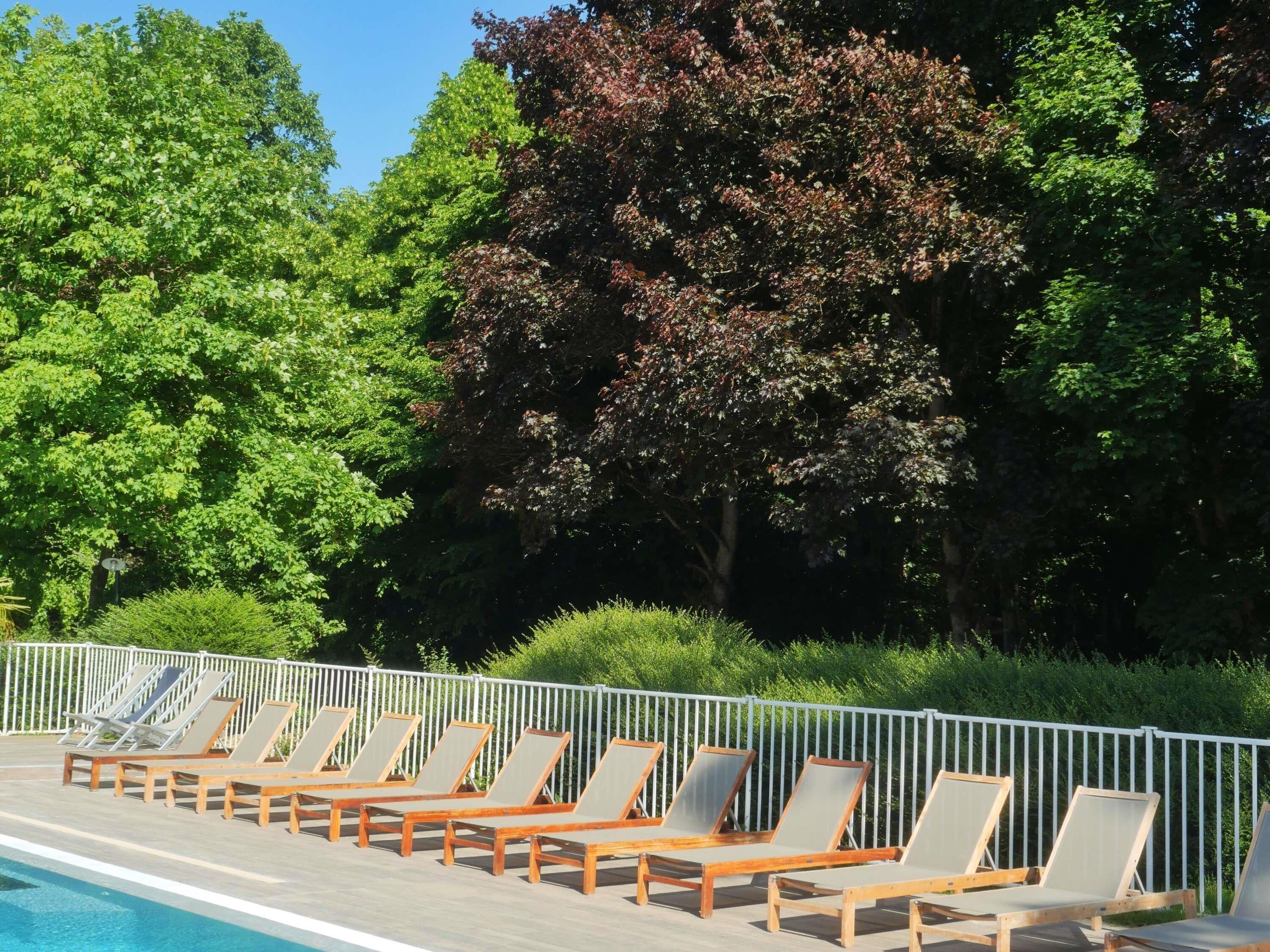 A poolside area with wooden lounge chairs aligned along the pool edge, a white metal fence, and dense greenery in the background.