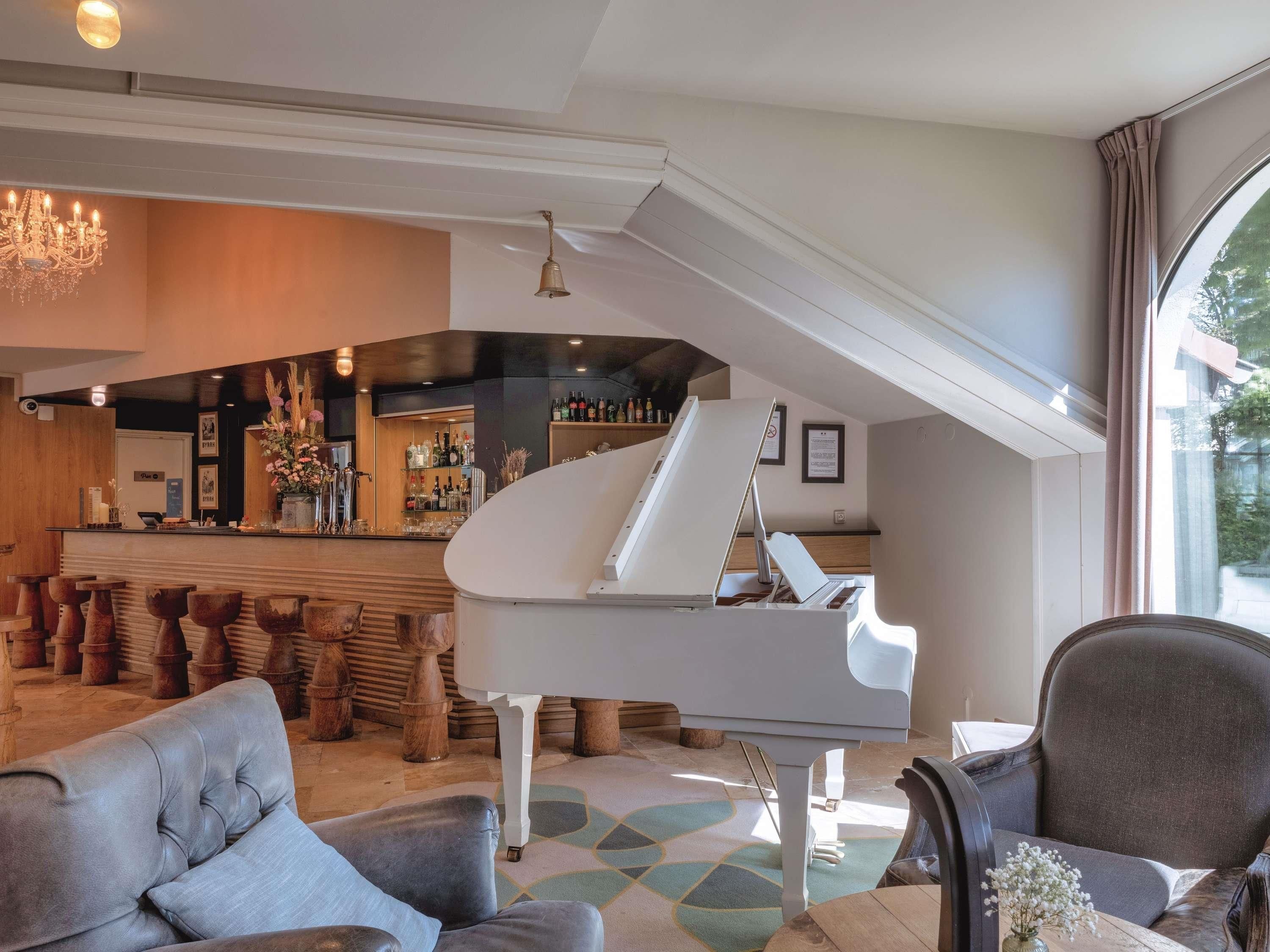 Un piano de cola, una zona de bar y un conjunto de asientos en una sala espaciosa con techo alto y ventanas amplias.
