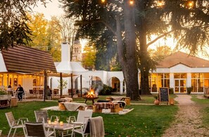 An outdoor dining area with tables and chairs, a fire pit, and a building with arched windows in the background. - Demeures de campagne Parc du Coudray – Mercure (Le Coudray-Montceaux)