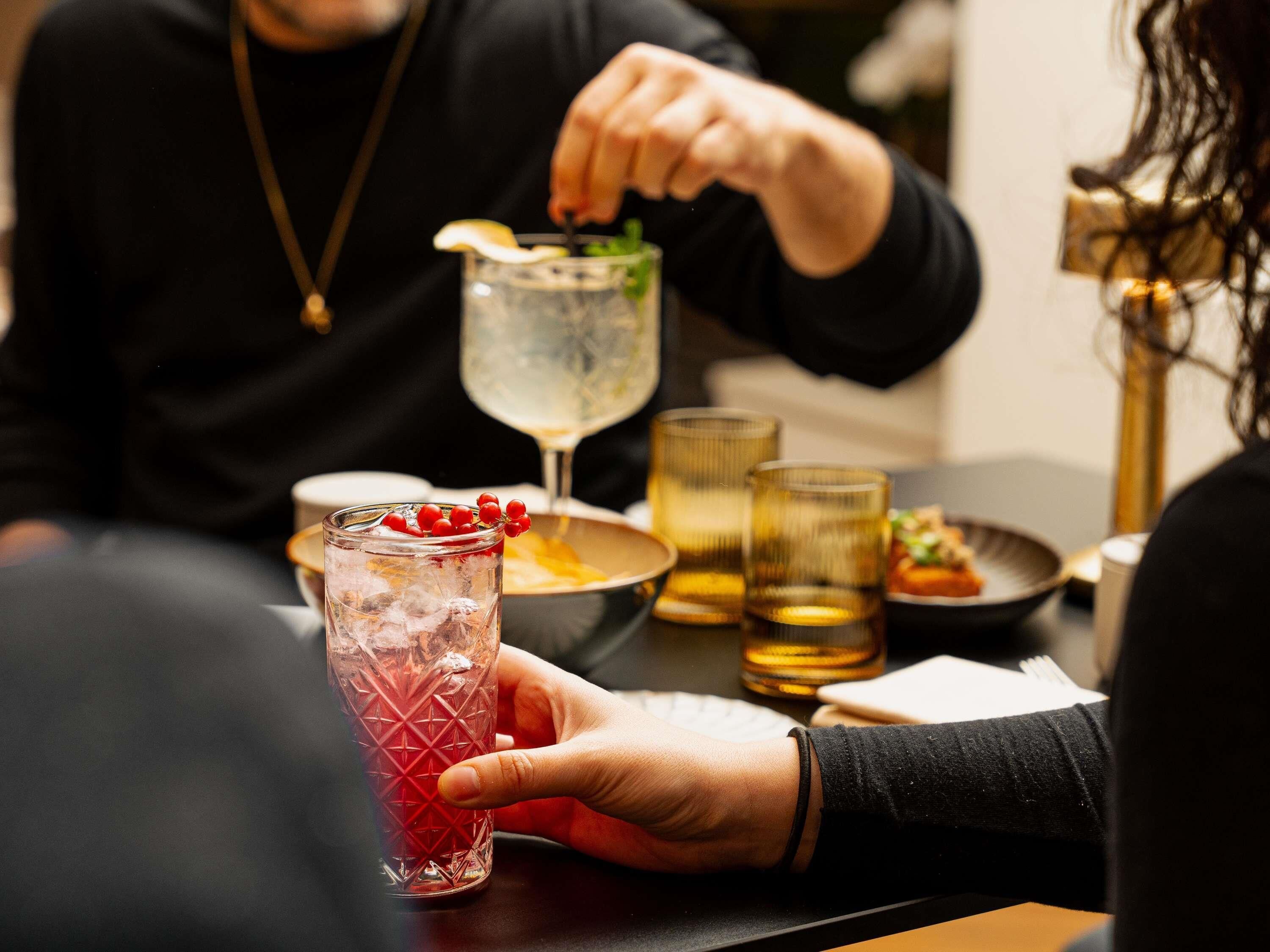 Una persona sostiene un vaso con una bebida roja, otro vaso con una bebida transparente y un plato de comida.