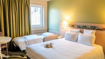 Une chambre d’hôtel avec deux lits simples, une petite table, une chaise, une table de chevet avec une lampe et une fenêtre avec des rideaux.