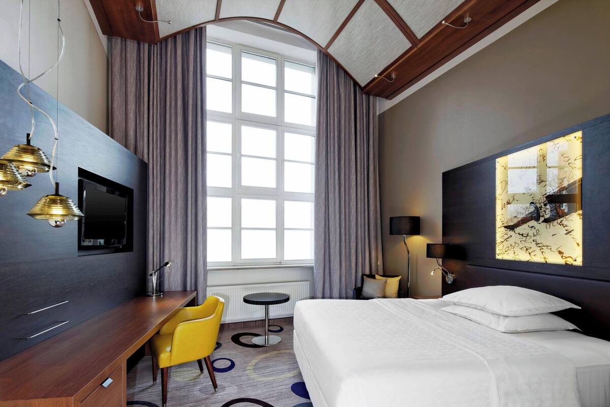A hotel room with a bed, a desk, a chair, a TV, and a large window with curtains.