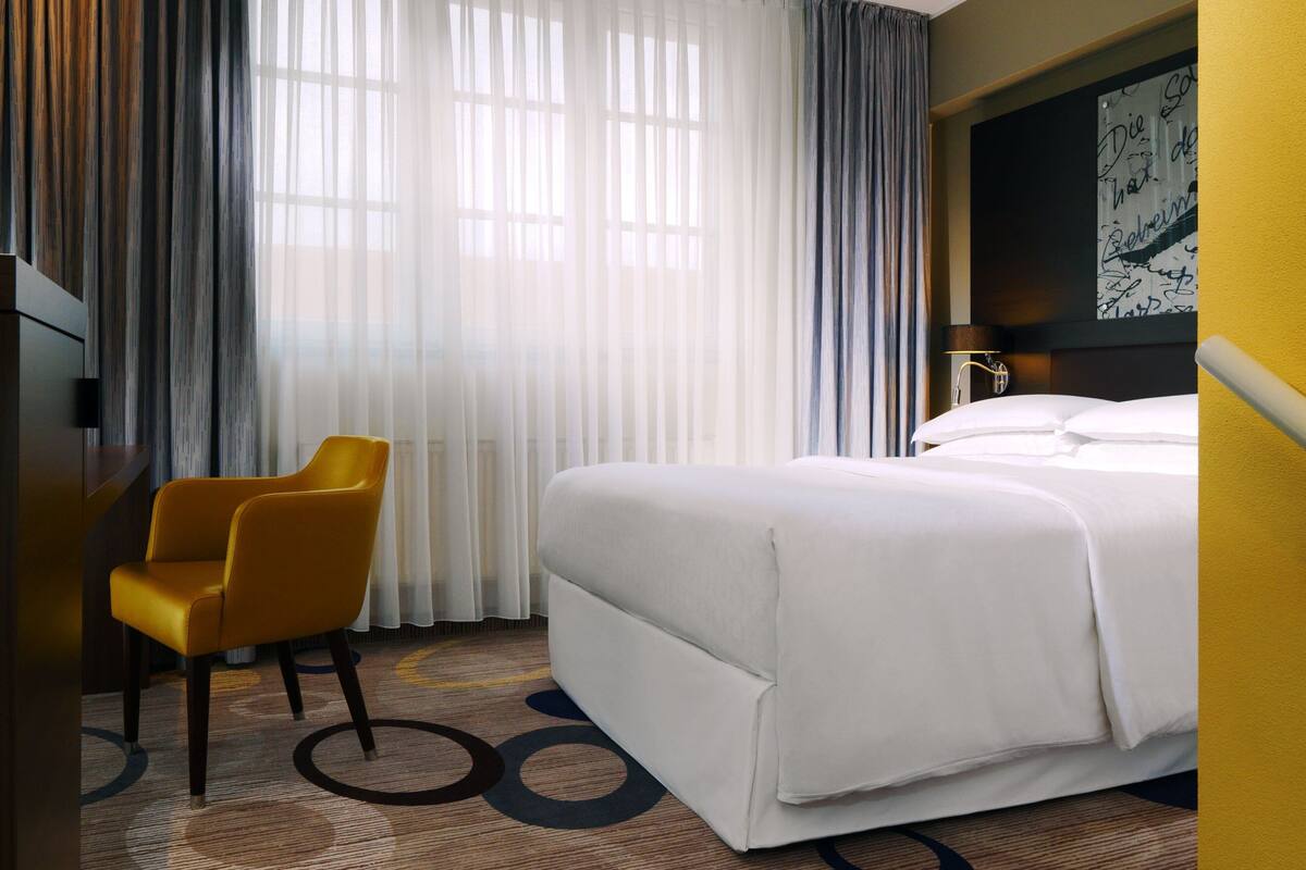 A hotel room with a bed, a chair, and a large window with curtains.