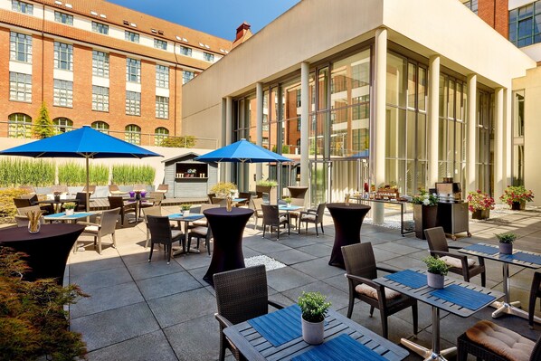 An outdoor patio area with tables and chairs, large windows, and a building in the background. - Sheraton Hannover Pelikan Hotel (Hannover)