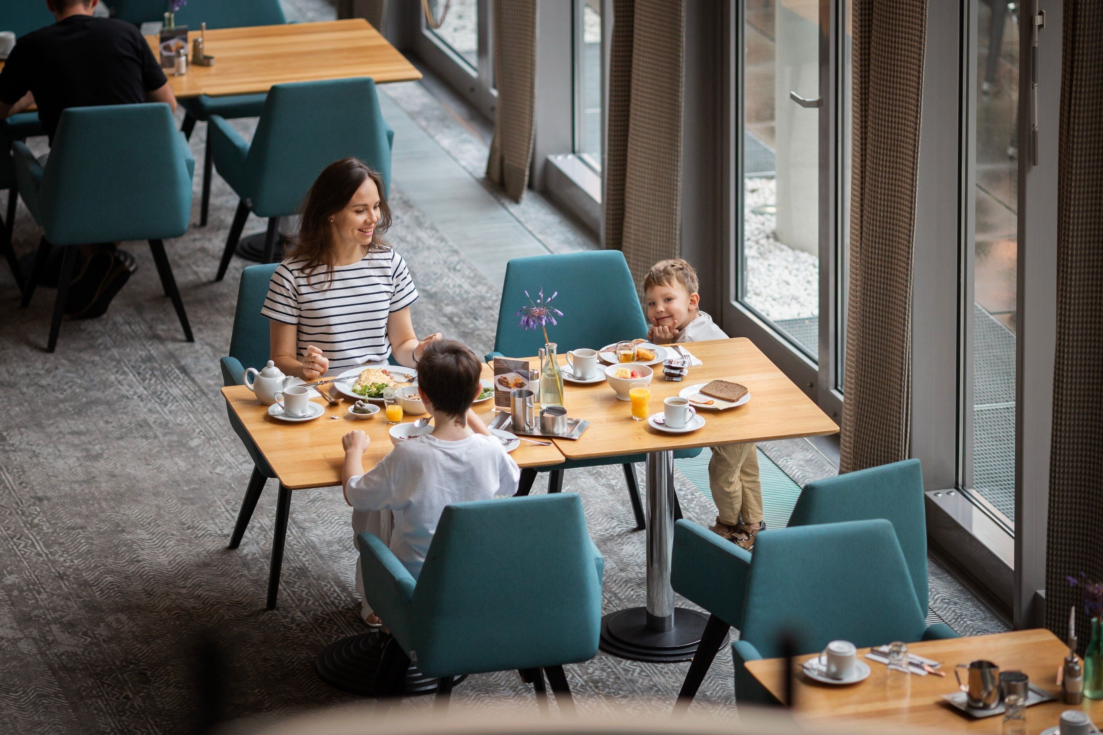 Een eetgedeelte met blauwgroene stoelen en houten tafels, met ontbijtartikelen en een vaas met bloemen.